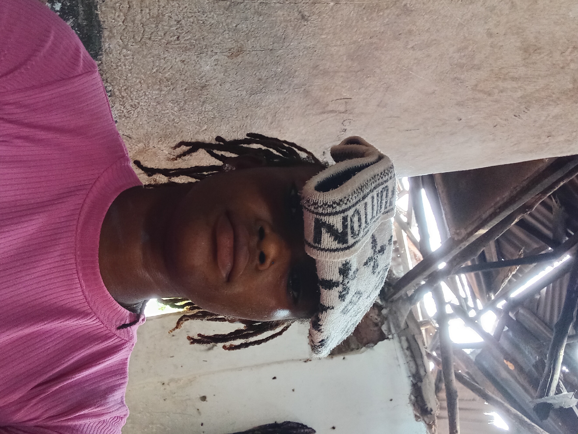 The image displays a person with dark skin and dreadlocks, captured in a close-up, slightly tilted portrait. The individual is wearing a bright pink, ribbed long-sleeved shirt and a white and black knitted beanie. The beanie features visible text, including the word "NOTION." Their face is directed towards the viewer, with a neutral expression, lips slightly parted.
The background reveals a structural interior, possibly a damaged or unfinished ceiling or roof. Exposed wooden beams form a crisscross pattern, through which bright sunlight streams, creating strong highlights and shadows. This suggests the photo was taken during the daytime. The walls or surrounding surfaces appear rough and light-colored. The overall impression of the setting is one of disrepair, construction, or a very rustic environment. The image is oriented on its side, making the person appear to be looking horizontally.