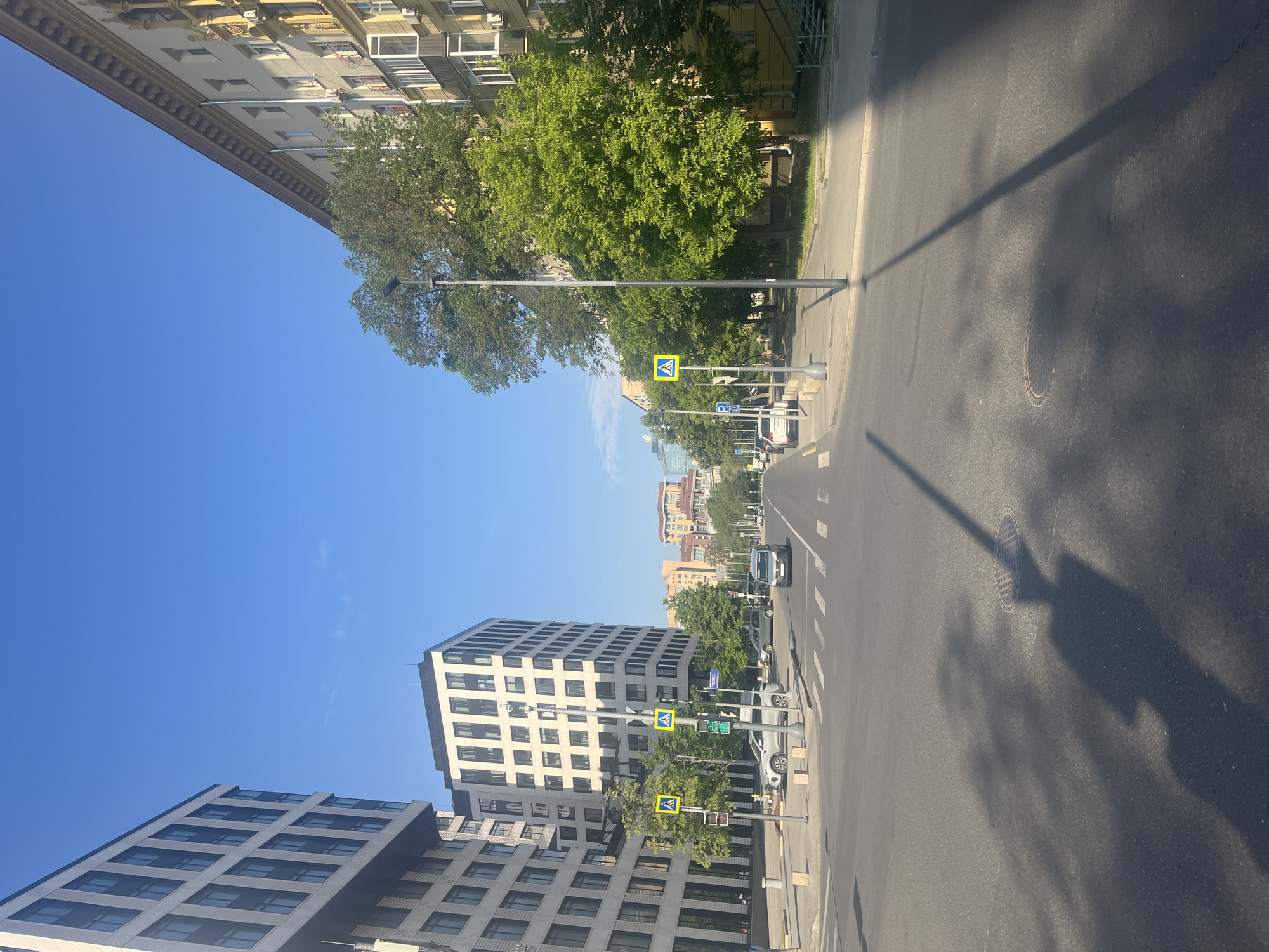 The image shows a street scene in Moscow, Russia, on a clear, sunny day. On the left, a modern white building with dark windows dominates the foreground. To its right is another, taller modern building, also with a grid of dark windows. Across the street, on the right side of the image, is an older, yellowish apartment building with balconies and ornate architectural details. Trees line the sidewalk in front of the older building.
The street itself is paved and appears to be a multi-lane road. Several cars are visible, including a silver SUV and a silver sedan in the foreground, and other vehicles further down the street. Pedestrian crossings are marked on the road. Yellow pedestrian crossing signs are posted on both sides of the street. Streetlights are also visible.
The sky is a vibrant blue with a few wispy clouds. The shadows cast by the buildings and streetlights suggest the sun is positioned to the right and slightly behind the viewer. The overall impression is of a well-maintained urban environment on a pleasant day.