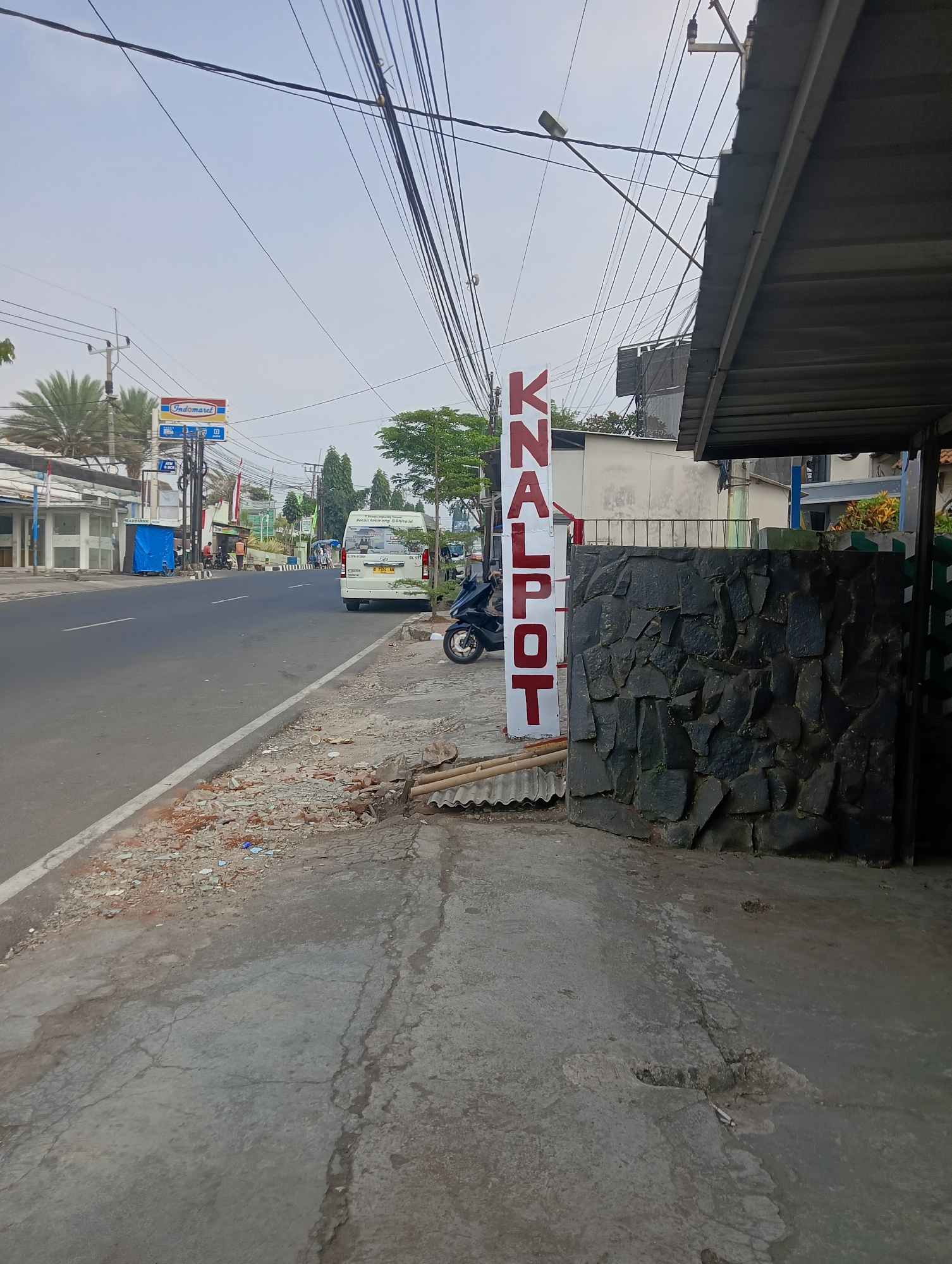 In Ciporang, Indonesia, a street scene unfolds under an overcast sky, dominated by a prominent vertical sign displaying "KNALPOT" in large red capital letters against a white background. To the right of this sign, a dark, irregularly shaped stone wall serves as a visual barrier. The foreground is a cracked concrete sidewalk, littered with debris and construction materials, including corrugated roofing sheets and scattered rubble. Beyond the sidewalk, a paved road runs horizontally, with a white line marking its edge. Vehicles are present on the road, including a white van with license plate B 1772 FU and a black motorcycle parked on the sidewalk to the left of the stone wall. On the left side of the road, a row of businesses lines the street, one of which has an "Indomaret" sign. Power lines crisscross the sky, supported by utility poles. The overall environment suggests a commercial area in a developing urban setting.