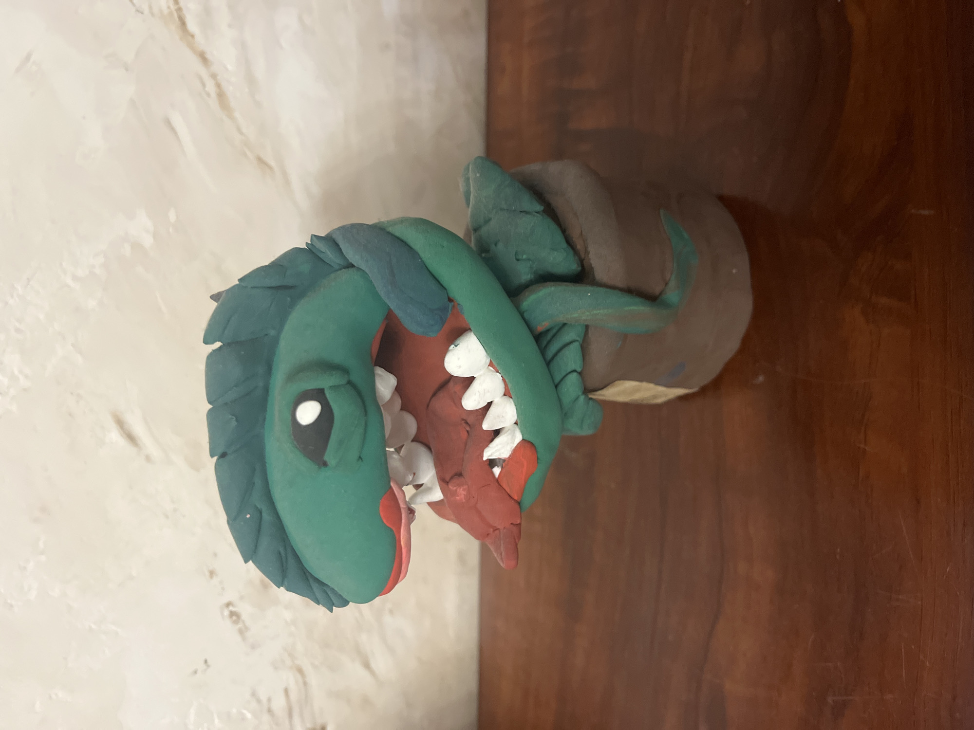 A handmade clay figurine of a green, plant-like creature with a large mouth full of white teeth sits in a brown pot on a wooden surface. The creature has a green, leaf-like top, a red mouth with a pink interior, and white teeth. Two green vine-like appendages extend from the creature and wrap around the pot. The background is a light-colored, textured wall. The image is taken from a slightly high angle, looking down at the figurine. No specific location in Vladimirovka, Russia can be determined from the image.