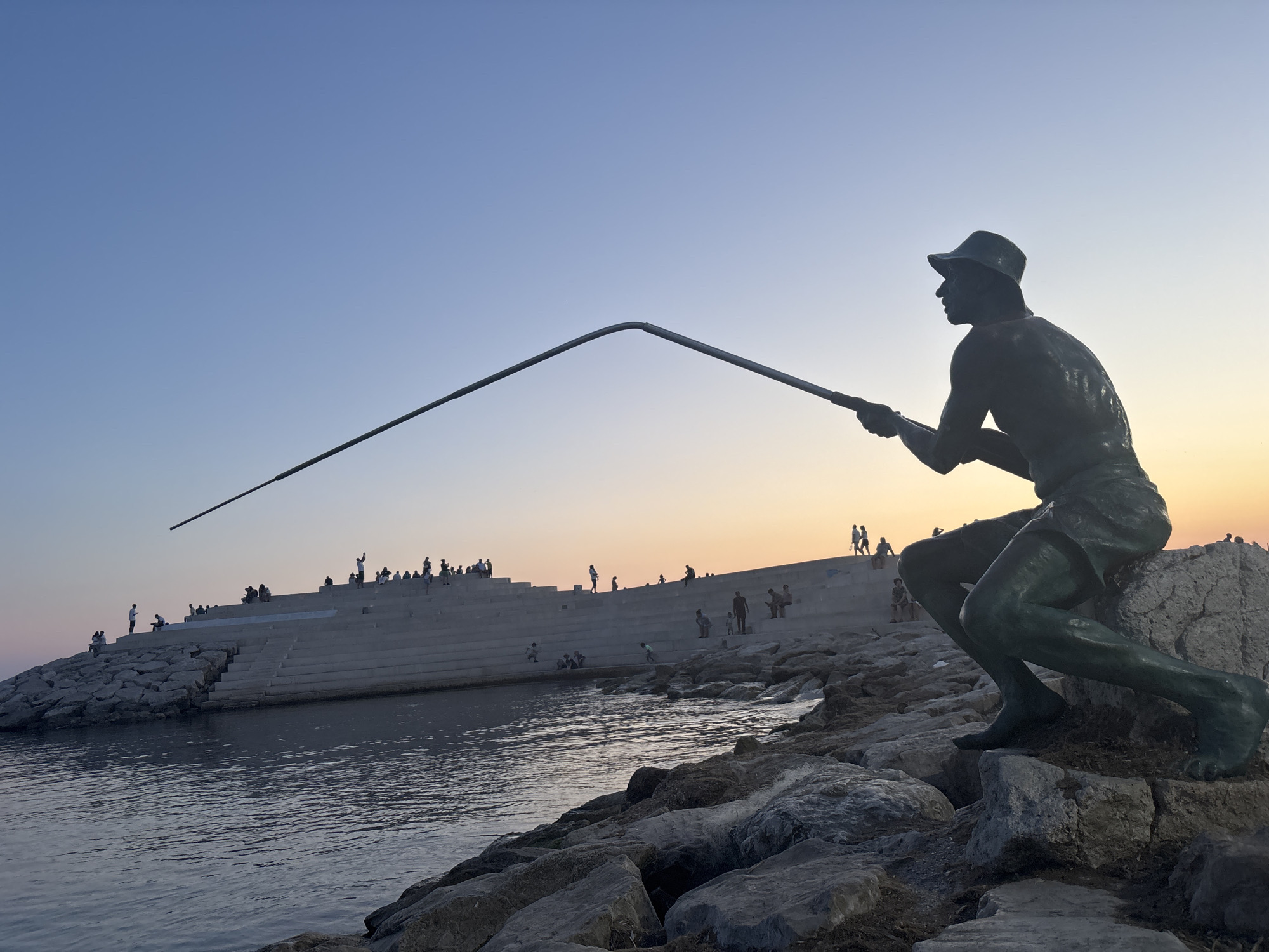 The image captures a bronze statue of a fisherman silhouetted against a clear blue sky with a soft orange glow near the horizon, indicative of sunset. The statue is positioned on a rocky shoreline, with its fishing rod extending upwards and to the left. In the background, a large, tiered concrete structure, possibly a promenade or amphitheater, is visible, populated by numerous small figures of people. The water in the foreground appears calm. The scene is set by the sea, with a breakwater made of large stones to the left of the statue. The location is identified as the "Statue of the Fisherman" in Bashkia Durrës, Albania.