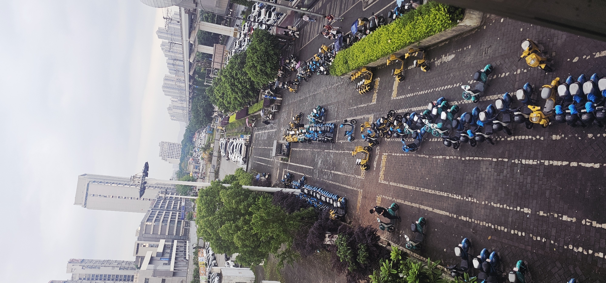 Here is a description of the image:
The image shows an aerial view of an area in the Chongqing High-tech Zone, China, filled with parked shared electric scooters and bicycles. The scene is oriented with buildings and a raised roadway visible in the background, indicating an urban setting. The foreground is dominated by rows of various electric scooters and bicycles, organized in designated parking areas marked by white lines on the paved surface. Scooters are predominantly in blue, white, and yellow. Some scooters are also parked in rows. Trees and landscaping are interspersed between parking areas, adding some greenery to the urban environment. The area seems to be an active transportation hub with numerous vehicles parked and likely used for local transportation. The overall atmosphere is urban, with a mix of transportation infrastructure and green spaces within the High-tech Zone.