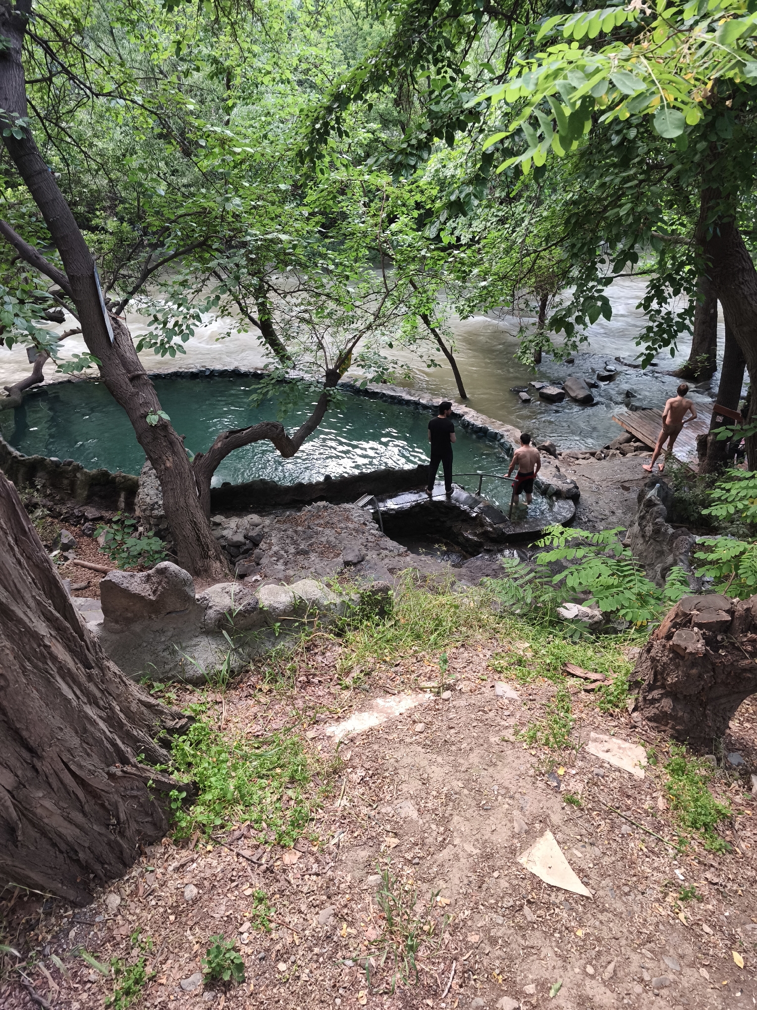 The image depicts an outdoor natural scene with a body of water that appears to be a river or stream, and a man-made pool. The pool, filled with clear turquoise water, is bordered by rocks and concrete structures. Several people are present. Three men are visible near the pool and the adjacent waterway. One man in black clothing stands on a concrete ledge overlooking the pool. Another man, shirtless and wearing red shorts, is standing in the shallow edge of the pool. A third man, also shirtless and wearing patterned shorts, is standing on a wooden platform near the flowing water. The environment is lush and green, with numerous trees and foliage surrounding the water features, suggesting a natural, possibly park-like setting. The ground in the foreground is covered with dirt, leaves, and scattered debris. The overall impression is of a secluded, natural bathing area. The location is identified as Yerevan, Armenia.