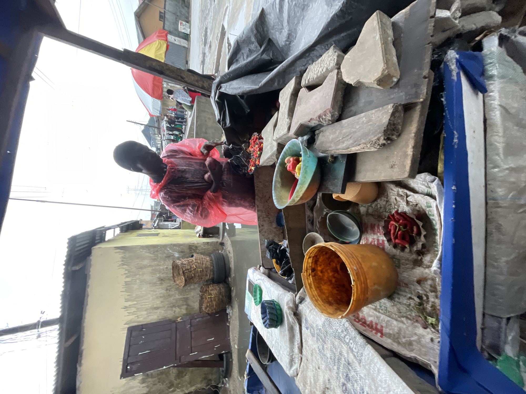 The image shows an outdoor market stall in Ado, Nigeria. A dark-skinned man wearing a red plastic poncho stands behind the stall, which is covered with a gray tarp. The stall displays items like stones, a blue and green bowl containing red and yellow items (possibly peppers and tomatoes), a brown plastic bucket, several other bowls, and a round container of red chilies. Woven baskets are visible next to a building with a closed brown wooden door, indicating the stall's proximity to residential structures. The sky is overcast, and the general environment suggests a modest urban setting.