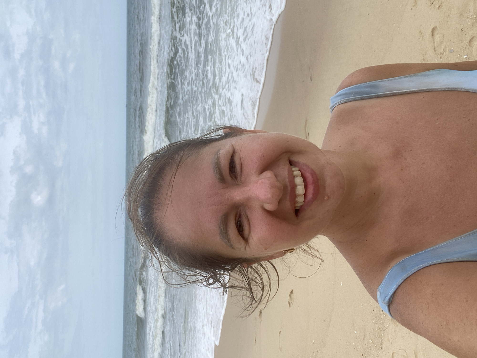 A middle-aged woman with light brown hair and fair skin is smiling at the camera in a selfie taken on a beach. She is wearing a light blue swimsuit. The ocean with moderate waves and a cloudy sky are visible behind her. The sand is light brown and appears damp in places, with a few footprints. The location is the city of Mũi Né, Vietnam.