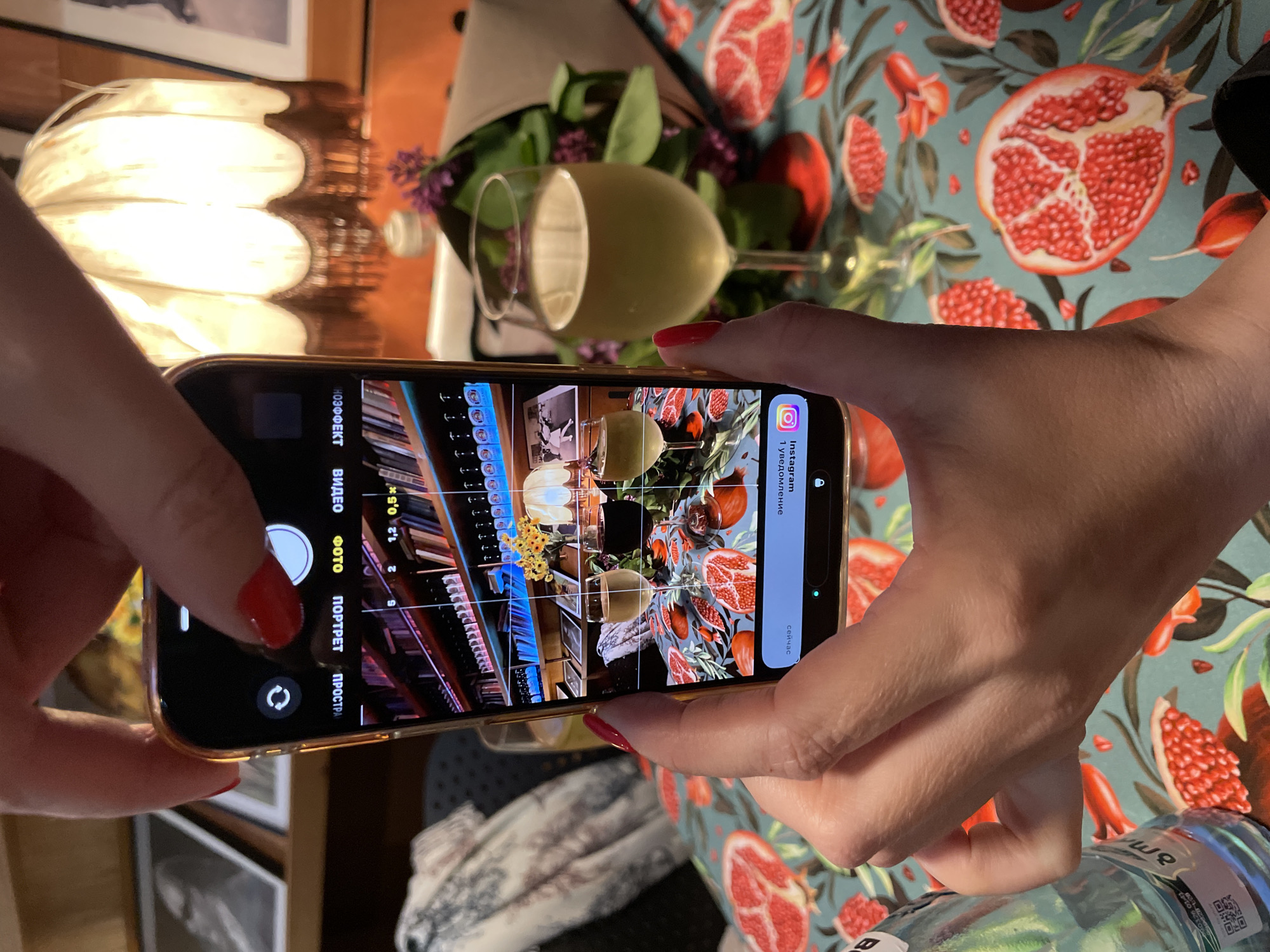 The scene depicts a person taking a photograph with a smartphone of a table setting. The table is covered with a patterned tablecloth featuring pomegranates. On the table, there is a wine glass filled with a light-colored liquid, likely wine or juice, and some flowers.
The phone screen shows a view of a bookshelf, wine bottles, and other objects, as well as the Instagram notification at the top.
The location is likely in Tbilisi, Georgia, given the Cyrillic script on the phone's screen, the Instagram notification, and other visual cues in the image.