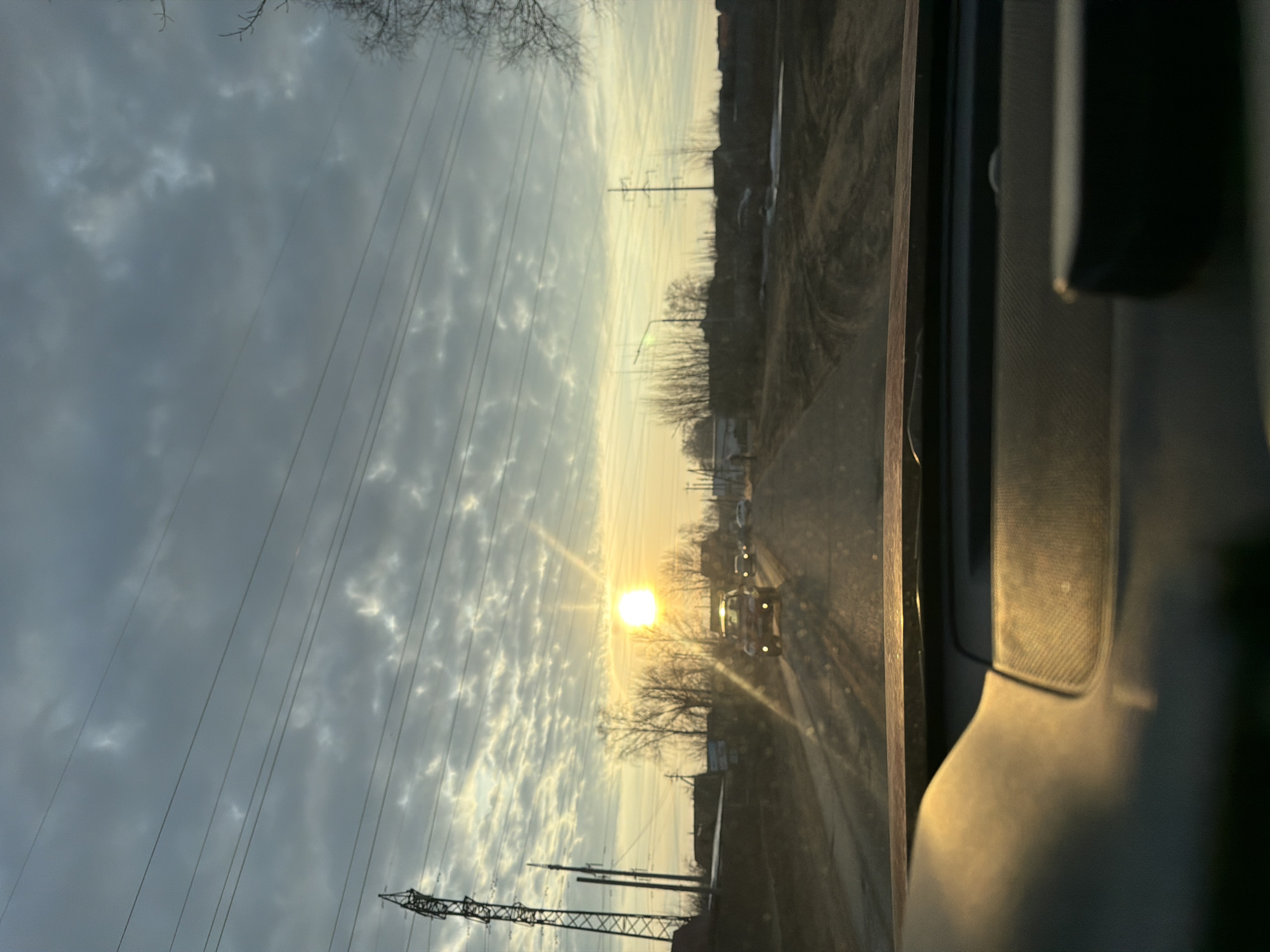The image depicts a view from inside a vehicle, looking out onto a road illuminated by the setting sun. Overhead, a sky filled with gray clouds is bisected by power lines. In the distance along the road, other vehicles are visible. The location is likely in Chaikovsky, Russia, evidenced by the presence of power lines and roadside structures typical of a suburban or rural Russian environment. The sun's position suggests either sunrise or sunset. The interior of the vehicle occupies the right side of the frame, partially obscuring the view.