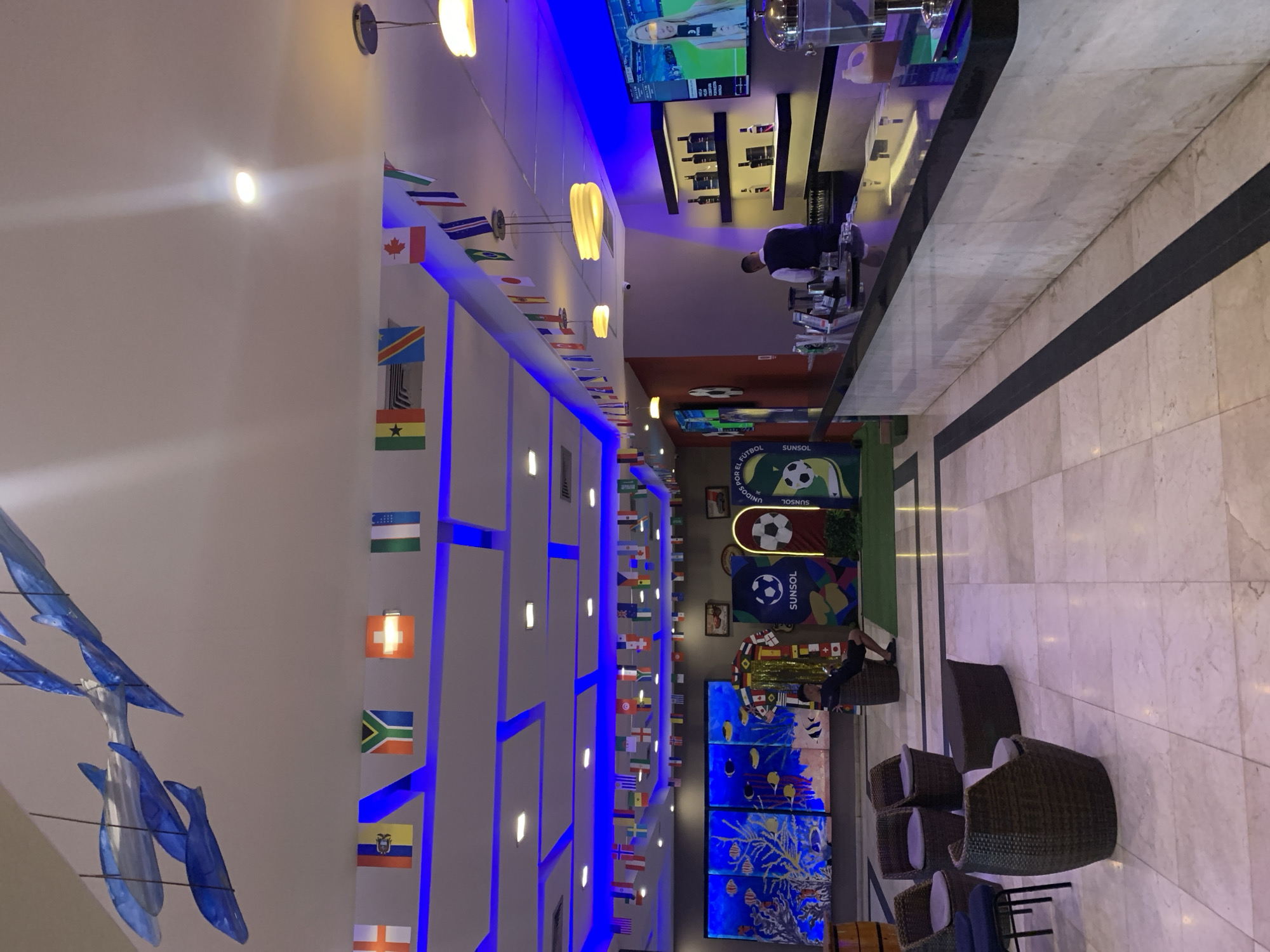 The image shows the interior of a venue decorated with numerous national flags strung across the ceiling. Blue lighting illuminates the space, highlighting a bar area with a bartender preparing drinks and a television displaying a sports broadcast. Seating areas with wicker chairs and tables are visible, along with a large mural depicting an underwater scene. Several football-themed decorations, including a large jersey and a soccer ball logo, are also present. The floor is tiled, and the ceiling features a modern design with recessed lighting. The overall ambiance suggests a sports bar or event space in El Tirano, Venezuela, likely set up for a tournament or celebration.