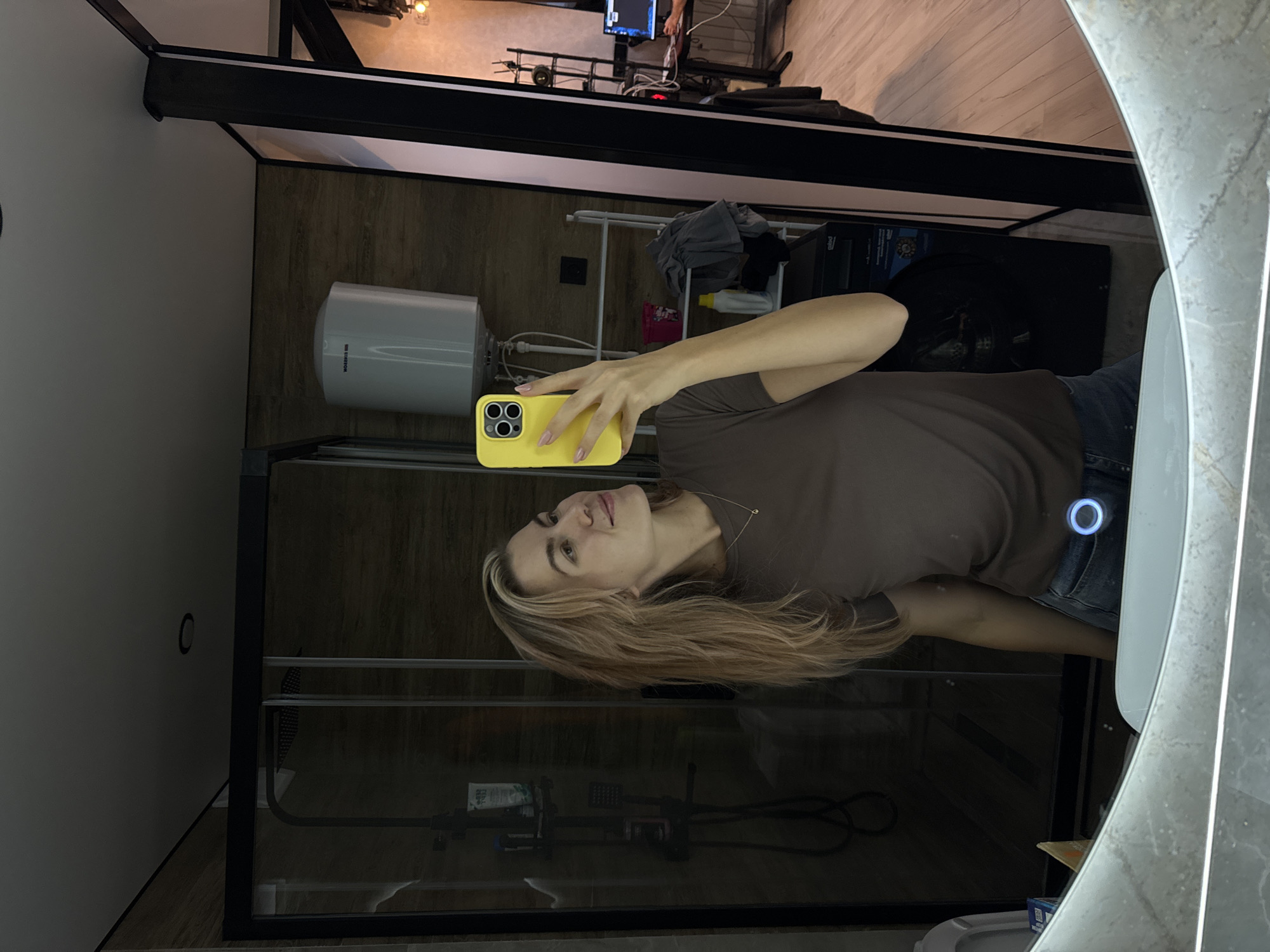 The image shows a woman taking a selfie in a bathroom. She is holding a yellow smartphone in her right hand, capturing her reflection in a large, rounded mirror with a marble-like frame. The bathroom features a shower enclosed by a dark-framed glass partition. Behind the woman and the mirror, there's a hot water tank mounted on the wall and a shelf unit with various items. The lighting in the room is a mix of natural and artificial. The location is indoors in a residential bathroom.