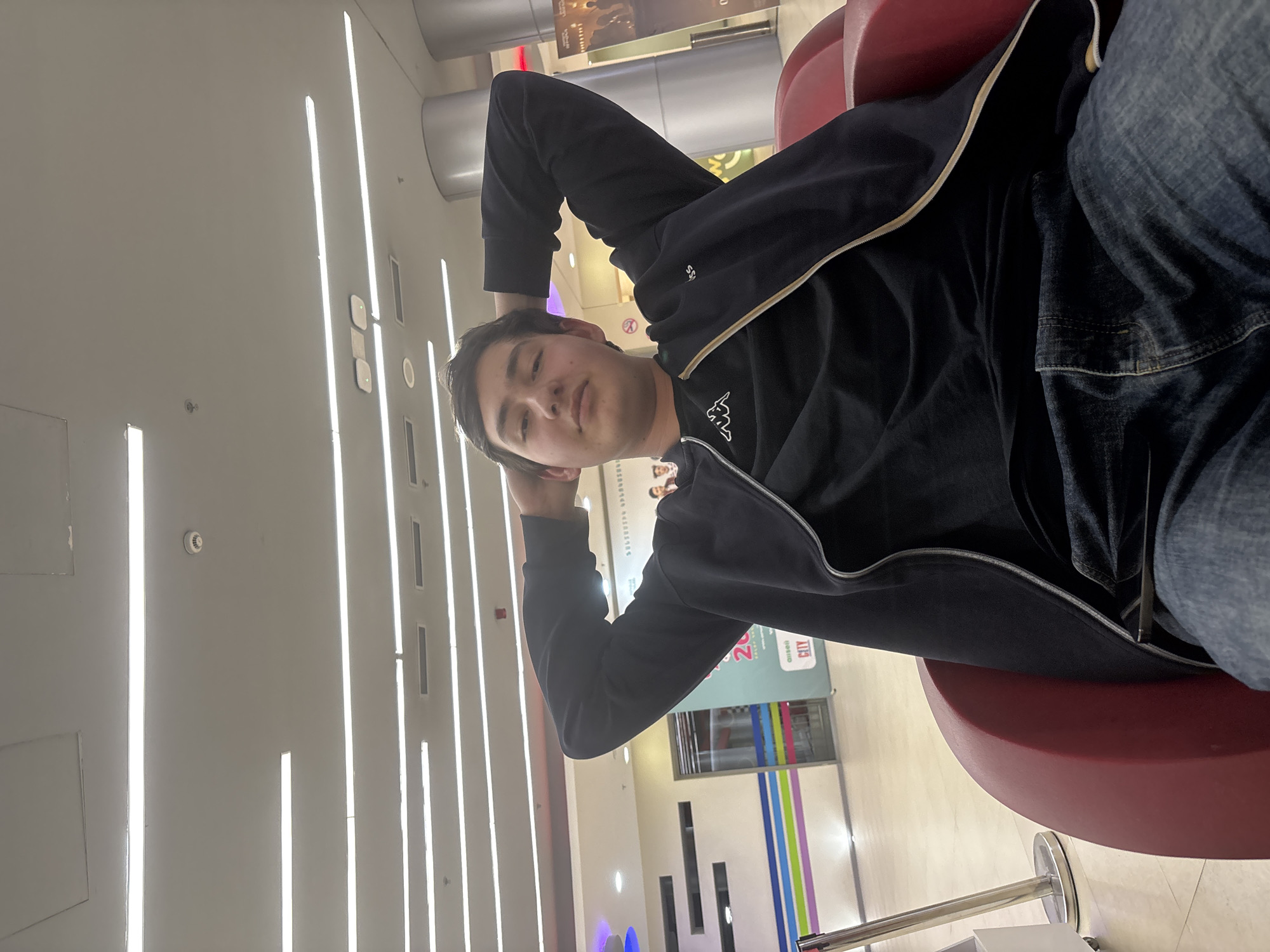 The image shows a young man resting in a red chair within a brightly lit indoor space. The walls feature vertical light strips and architectural details, suggesting a modern building interior. The man is wearing jeans and a dark zip-up jacket with a Kappa logo. He has his hands behind his head, appearing relaxed. Based on available details, it is not possible to determine the specific location within Yerevan, Armenia. However, the setting resembles a shopping mall or transportation hub.