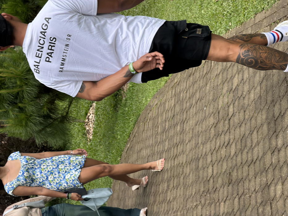 A man wearing a white t-shirt with "BALENCIAGA PARIS" printed on the back, black shorts, and white shoes stands on a paved path. He has tattoos on his leg. A woman in a blue and white floral dress and beige sandals stands near him, holding a purse. Part of a third person's green pants and shoes are visible. Green grass and foliage are on either side of the path.