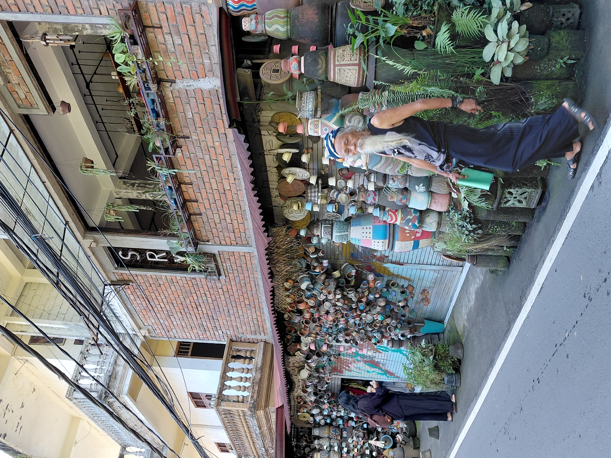 In Ubud District, Indonesia, a man with a long white beard, wearing a black tank top, black pants, and a blue and white bandana, walks past a storefront filled with decorative pottery. The storefront displays a wide assortment of ceramic pots, bowls, and other decorative items, many of which are stacked and arranged both inside and outside the shop. A woman in a black outfit and a brown backpack stands near the entrance of the shop, looking at her phone. The building behind the shop is constructed of brick with balconies and white balustrades. Overhead, a tangle of electrical wires spans the street. The scene appears to be a street-level view of a commercial area specializing in artisanal crafts.