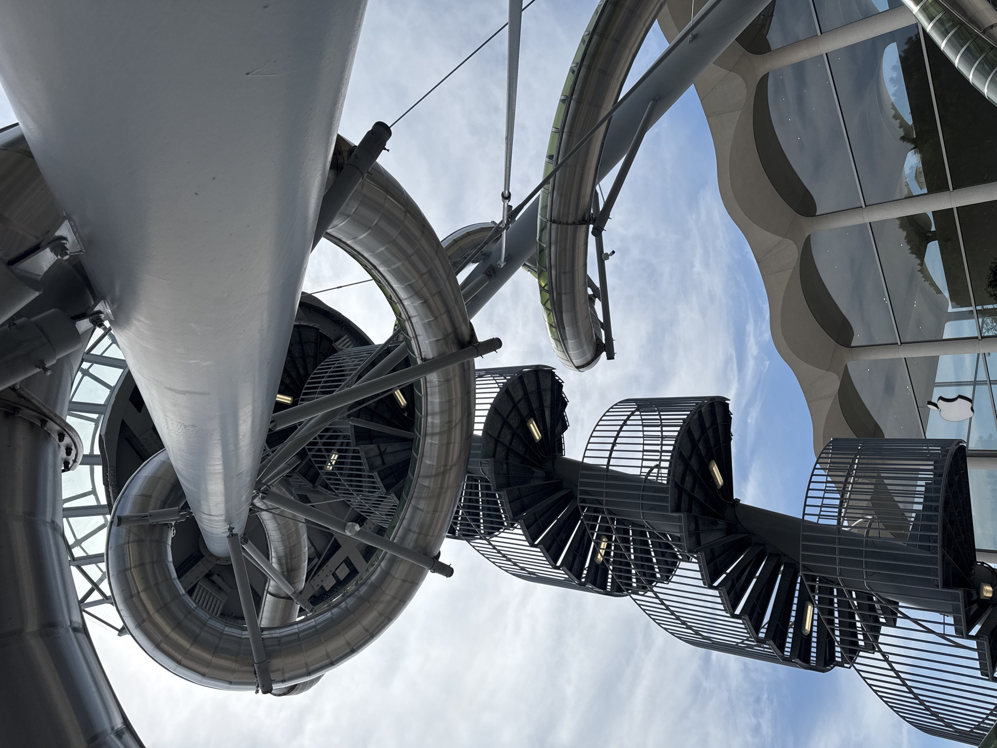 An exterior low-angle shot captures a segment of a contemporary architectural structure in Aventura, Florida, United States of America, under a partly cloudy sky. The primary elements are a large, metallic, tubular slide system and a black spiral staircase. The slide is a prominent, silver, curved structure that appears to be made of metal, with sections that are translucent green. It is supported by a complex network of grey metal beams and struts.
To the left of the slide, a black spiral staircase with metal railings ascends, featuring a central support column and several landings. Each landing is illuminated by a yellow light fixture. The staircase is intricate and appears to be made of dark metal.
In the lower right portion of the image, a building with a unique, wavy, cream-colored facade is visible. Large glass windows reflect the sky and some trees. A distinct white Apple logo is affixed to the glass facade, indicating the building is an Apple Store. The overall scene suggests a public space or a unique piece of urban infrastructure, possibly part of a park or recreational area adjacent to the retail store. No people are visible in the frame.