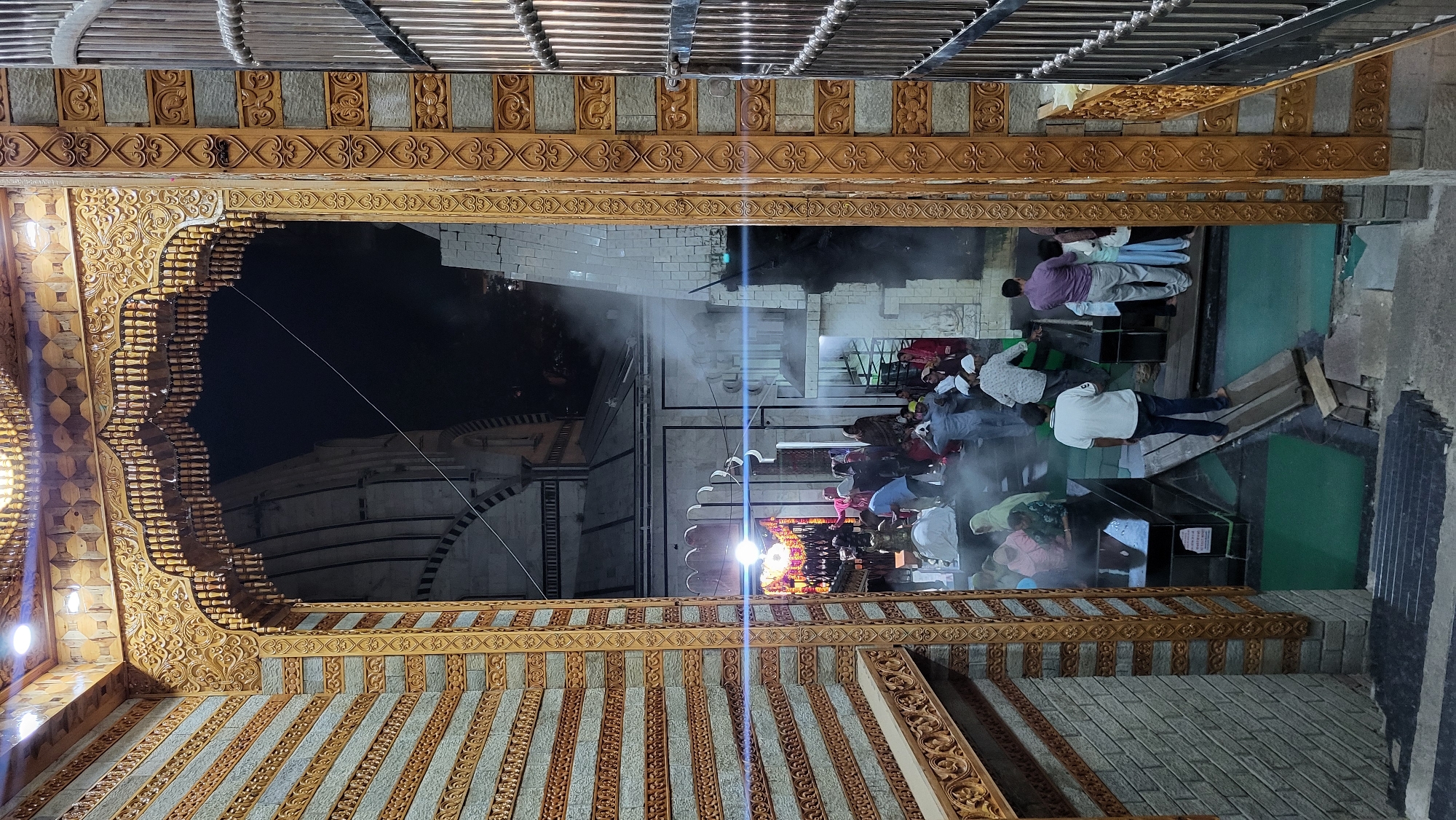 The image depicts a nighttime scene viewed through an ornate wooden archway, revealing a gathering of people outside a large building, presumably in Kasol, India. The prominent archway, serving as a frame, is richly decorated with intricate carvings featuring floral motifs, geometric patterns, and numerous small wooden spindles. Above the archway, a patterned ceiling with carved wooden strips and a metallic railing are visible.
Beyond the arch, a crowd of people is gathered. Some individuals are standing, while others are seated on the ground, which features sections of green matting or carpeting. Several men in the gathering are wearing skullcaps, suggesting a religious or community event, possibly within a Muslim context. The activity appears to be a communal congregation.
In the background, a substantial building with a distinct dome-like or tiered roof structure stands against the dark night sky. Its exterior walls are light-colored, possibly white brick or stone. The scene is illuminated by artificial lights, with a bright light source creating a prominent lens flare. A faint haze or mist, possibly smoke or atmospheric vapor, is visible throughout the area, illuminated by the lights. On the ground level within the gathering, there are some open cubby-like structures and wooden planks. A small, illegible sign can be seen on one of these structures. The setting conveys a sense of a peaceful evening assembly at a place of cultural or religious importance.