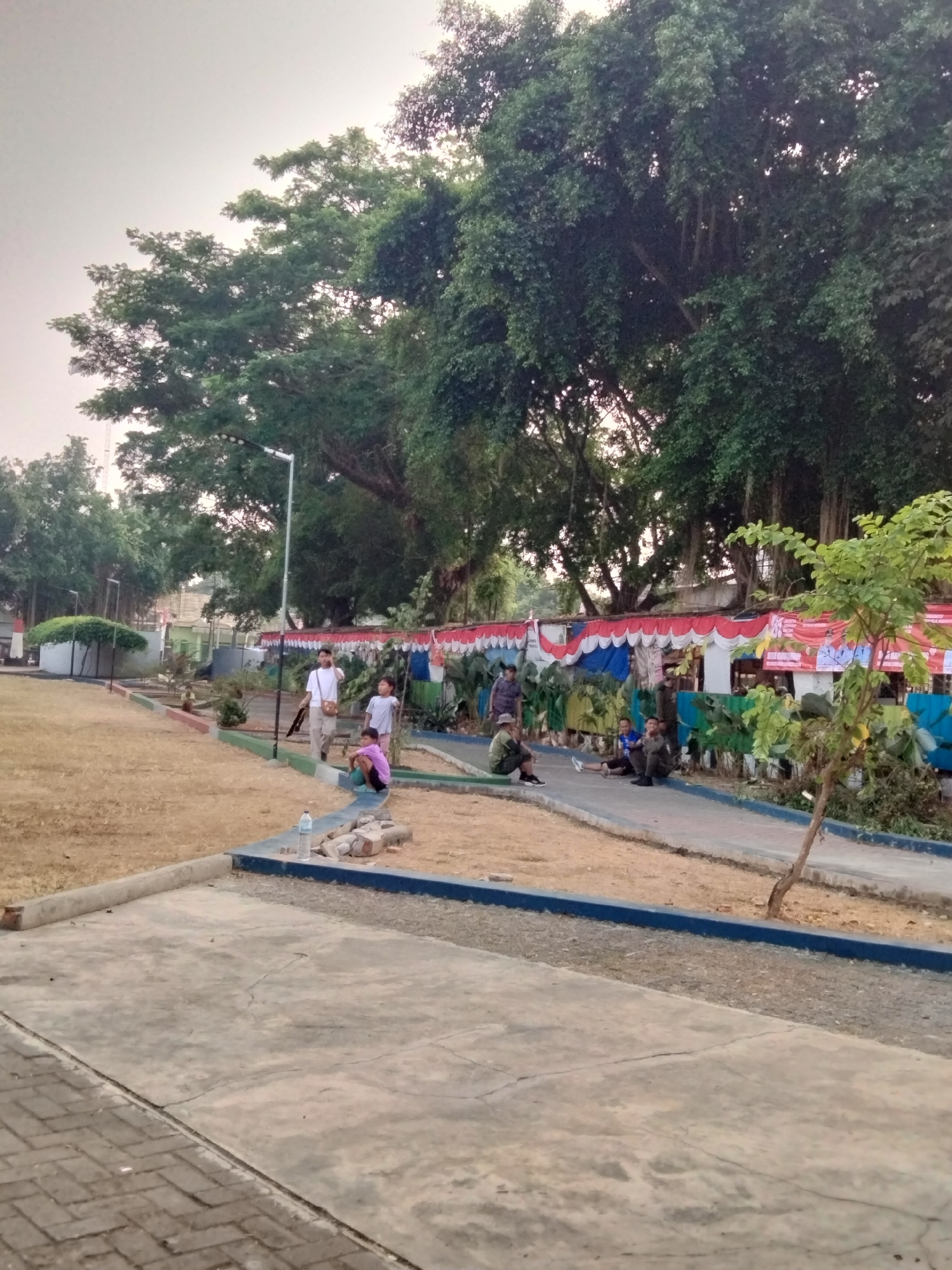 In Cariu, Indonesia, a group of people are gathered in an outdoor public space, possibly a park or recreational area, near a street. The environment features a prominent large tree with a thick trunk and dense foliage dominating the background, suggesting a tropical setting. The ground is a mix of dry, light brown dirt and paved concrete pathways. A decorative element, likely a temporary setup for an event, is visible along a fence line, featuring a canopy with red, white, and blue fabric.
Several individuals are present. In the foreground, a person sits on a low concrete barrier, facing away from the camera. To their left, a bottle of water and some scattered bricks lie on the ground. Further back, a man in a white t-shirt and beige shorts walks towards the camera, holding a bag. Beside him, two children, one in a white t-shirt and the other in a pink top, are also present. In the middle ground, a line of people are seated or leaning against a low wall in front of the decorative canopy. Some appear to be conversing. Another person stands near a lamppost in the left-center of the image. The overall lighting suggests it is daytime, possibly late afternoon or early morning, given the soft light and overcast sky.