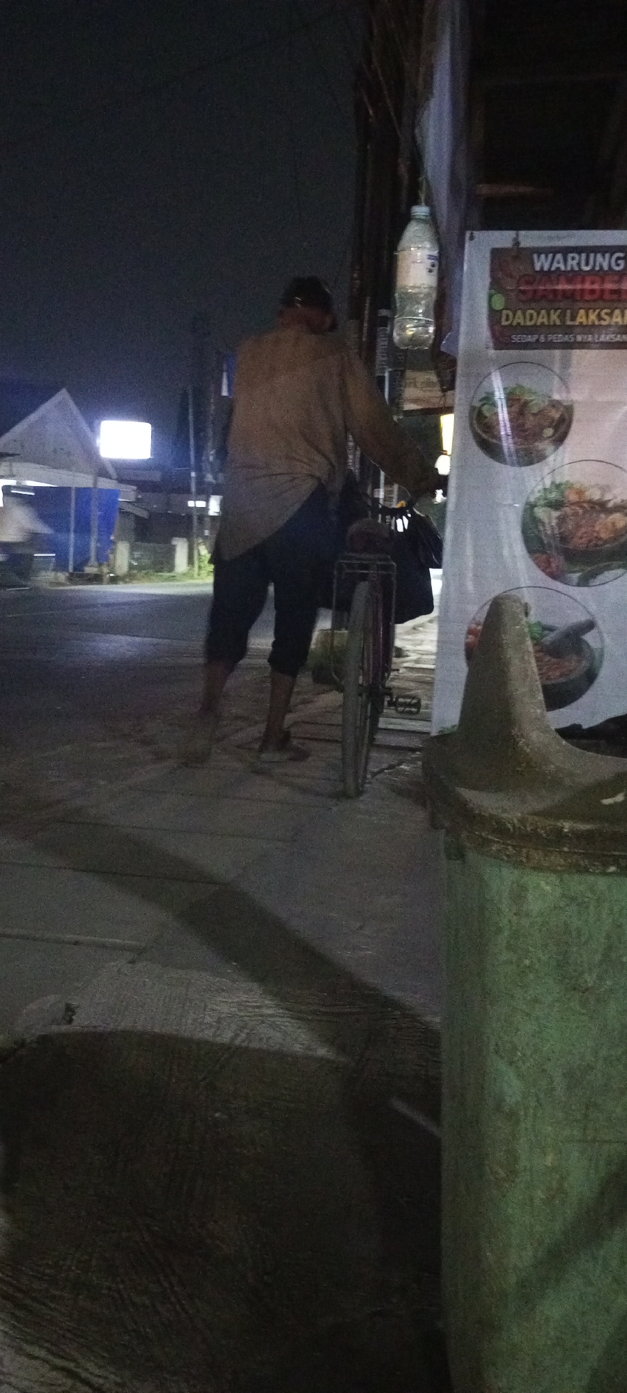 In the city of Setu, Indonesia, a person is seen walking alongside a bicycle at night. The individual, wearing a tan long-sleeved shirt and dark pants, appears to be pushing the bicycle, which has a dark bag attached to the rear. In the background, a white structure with a brightly lit rectangular sign is visible, along with some blue tarps. To the right of the frame, a banner for "WARUNG SAMBEL DADAK LAKSA" displays images of food. A green trash receptacle is situated in the foreground. The ground is paved, with shadows cast by the streetlights and surrounding objects.