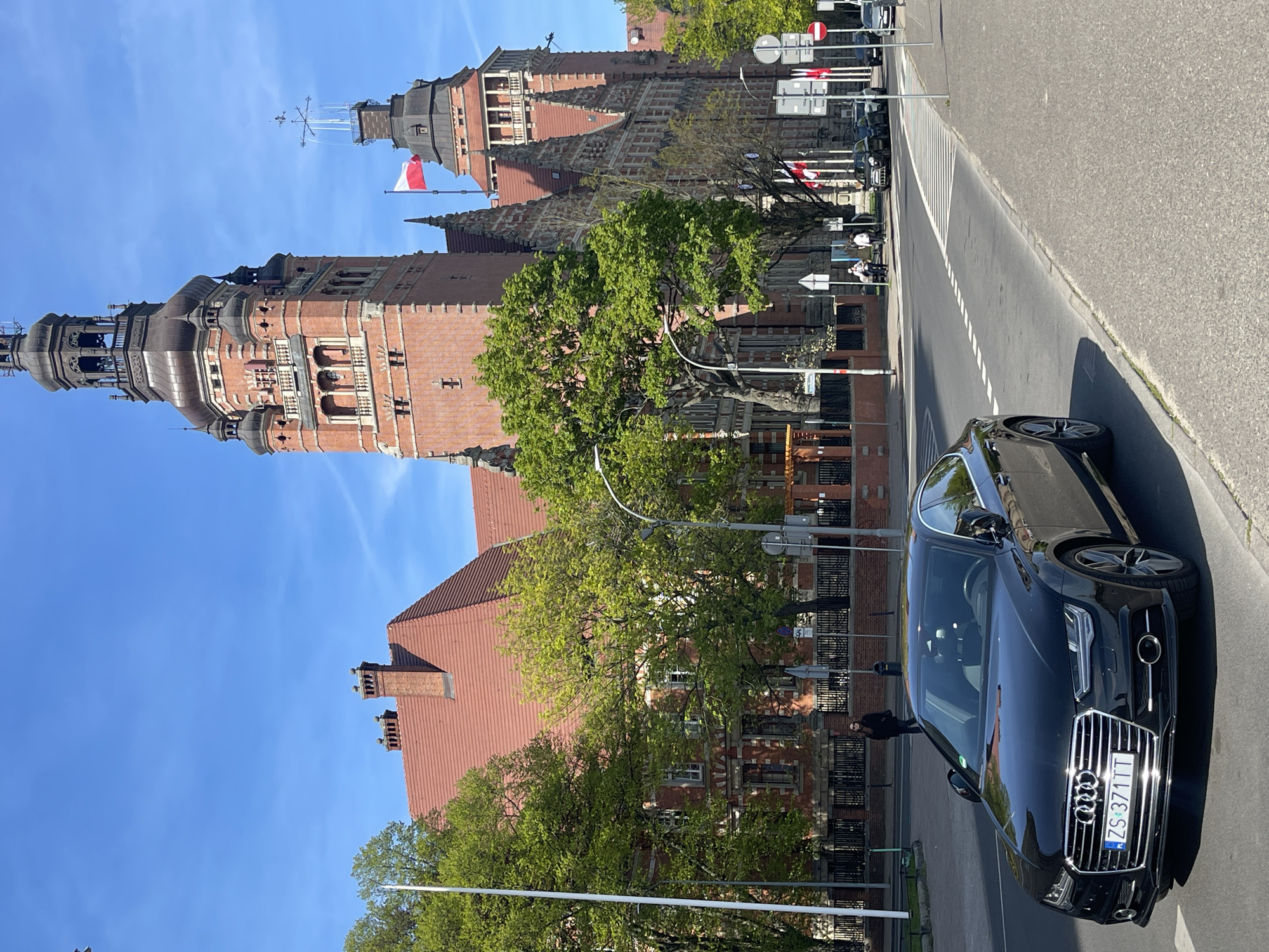 This image captures a vibrant street scene in Szczecin, Poland, under a clear blue sky on a sunny day, likely late morning or early afternoon given the bright lighting and shadows. The perspective is slightly upward, with a grand, historic building dominating the left side of the frame and a modern car occupying the lower right foreground.
The prominent structure on the left is an elaborate brick building featuring multiple towers with intricate detailing and red tiled roofs. Its architecture suggests a significant civic or academic institution. One of its towers, topped with a weather vane or mast, proudly flies a red and white Polish flag. To its left, a taller, more ornate tower with a dark, possibly copper-clad, dome-like roof extends higher into the blue sky. Lush green trees are interspersed along the front and mid-ground, partially obscuring the lower sections of the buildings. Another red-tiled roof with chimneys is visible behind the trees on the right side of the main building.
On the right side of the image, an asphalt road stretches into the distance, bordered by a sidewalk. White dashed lines mark the road, and a zebra crossing is visible further down. A sleek, black Audi car (likely an A7 model) is parked or moving slowly on the right side of the road, its front grille with the prominent Audi logo and license plate clearly visible. The license plate reads "ZS 371TT," indicating its registration in Szczecin. Several blurred figures of people can be seen walking or standing on the sidewalk in the mid-ground, near the crosswalk and trees, with one person more distinctly visible behind the car. Traffic signs are also scattered along the sidewalk, including a "no entry" sign.