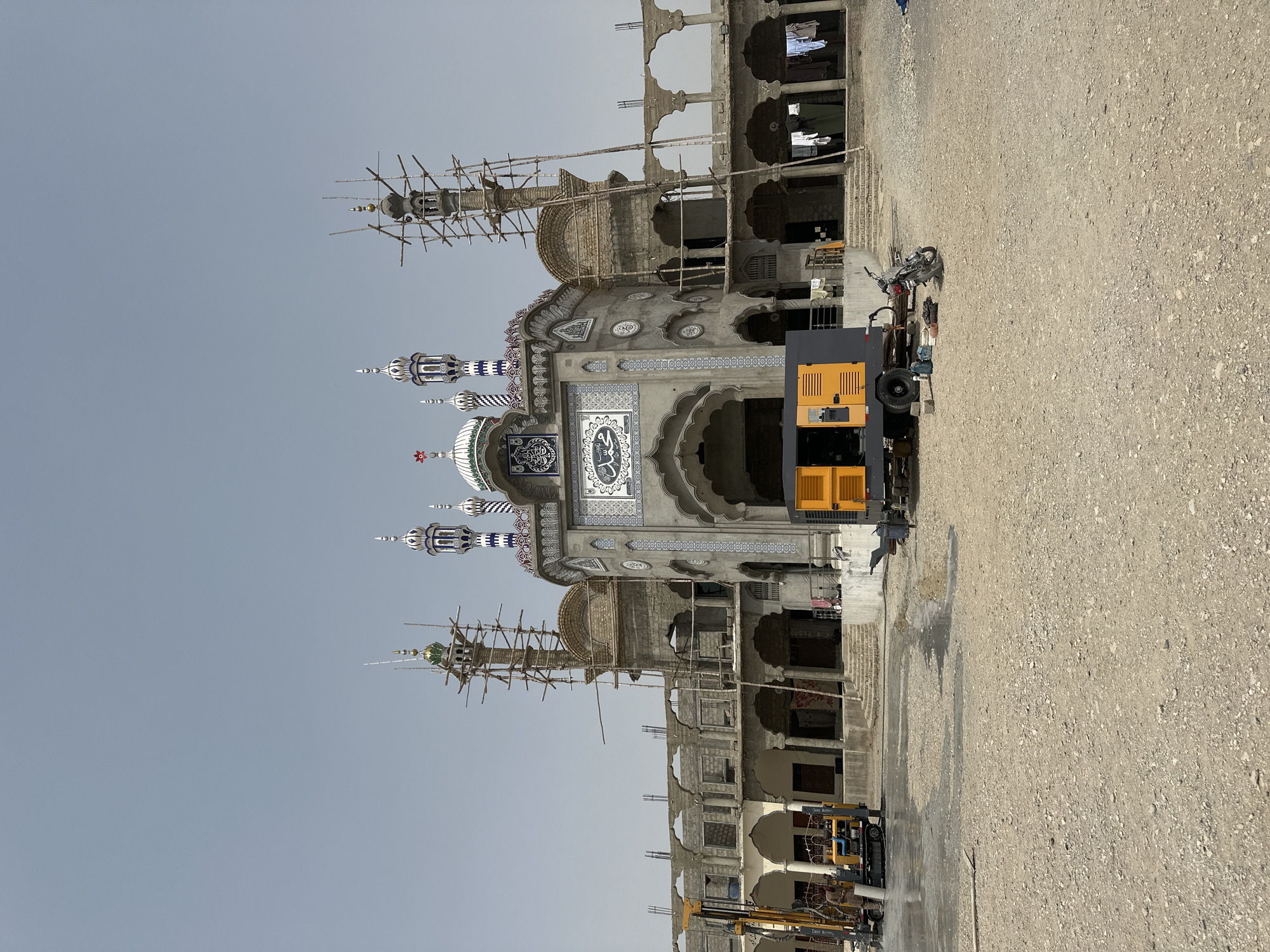 The image depicts a construction site for a mosque in the city of Khuzdar, Pakistan. The mosque, an Islamic religious building, is in the process of being built, with scaffolding visible around some of its structures. The facade of the central portion of the mosque is decorated with blue and white tile work, and Arabic calligraphy. A clock is prominently displayed on the facade, with Arabic numerals. Several minarets, characteristic features of mosques, are either partially completed or already decorated.
In the foreground of the scene, construction equipment is present, including a yellow drilling rig with a person standing next to it, and a large orange generator. A motorcycle is parked nearby, along with some tools and containers. The ground is a dusty, unpaved surface, typical of a construction area. In the background, the partially constructed building's colonnades and arches are visible, suggesting a larger complex. Laundry is hanging on lines in the background, indicating nearby habitation. The sky is clear and overcast, suggesting a possibly warm day. The overall scene conveys a sense of ongoing construction and development.