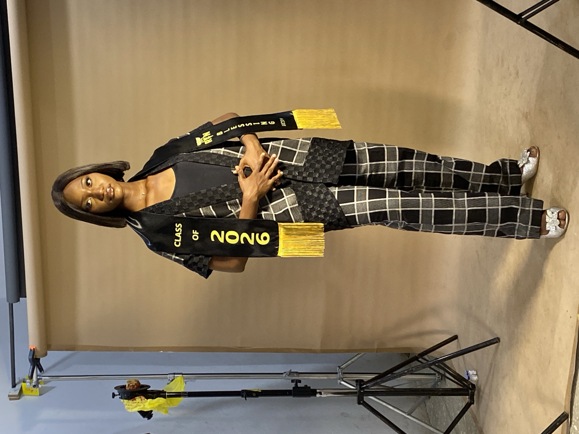 A full-length studio shot shows a dark-skinned young woman standing in front of a tan seamless paper backdrop. She is wearing a black tank top with a black and white checkered blazer and matching pants. Around her neck is a black graduation stole with "CLASS OF 2026" and "BLESSING" printed in yellow. She wears silver sandals. Her hands are clasped in front of her. Studio lighting equipment is visible in the foreground. The image does not depict the location in Gura Top, Nigeria, but appears to be a studio setting.