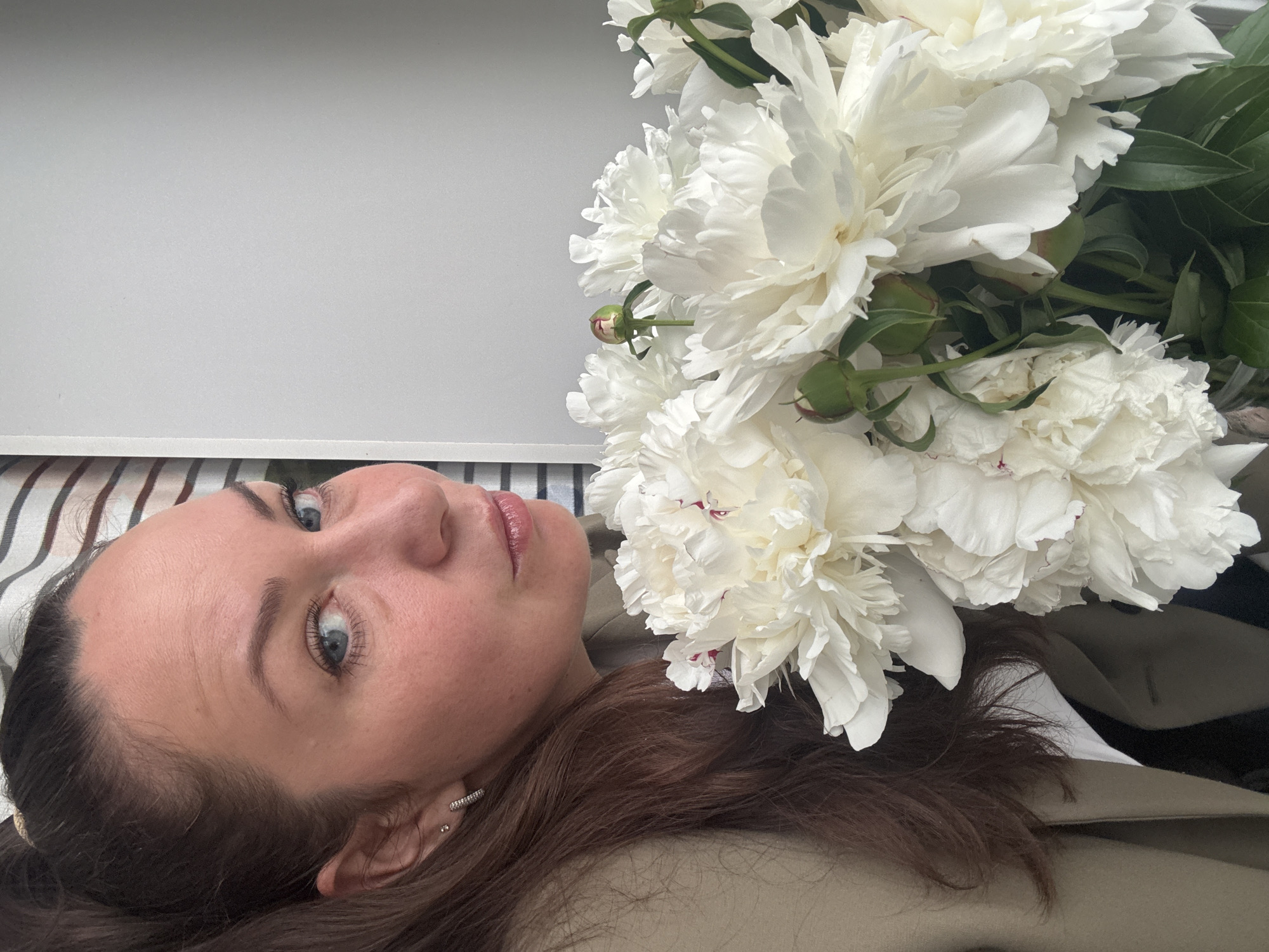 The image, presented in a landscape orientation, features a woman and a large bouquet of white peonies. The woman's head and upper torso are positioned on the left side of the frame, oriented on her side. She has fair skin, long brown hair flowing around her, and striking blue eyes that gaze directly at the viewer with a neutral and calm expression. Multiple small, sparkly ear piercings are visible on her left ear. She appears to be wearing a light-colored, possibly beige, jacket or coat. Her head rests on a striped surface, likely a pillow or bedding, visible at the bottom left.
Filling the right and upper portions of the image is a large, opulent bouquet of white peonies. The flowers are in full bloom, showcasing their numerous soft, ruffled petals. Some of the central petals exhibit subtle reddish or pinkish accents. Green foliage and a few unopened buds are visible among the blossoms. The upper part of the background is a plain, light grey surface, consistent with an indoor wall or flat object.
The scene conveys a serene and intimate moment, possibly a self-portrait, with the woman posed alongside the beautiful flowers. The lighting is bright and even, suggesting a daytime indoor setting. There is no visible text within the image. While the image is from Chaikovsky, Russia, no specific visual cues in the photograph indicate this location.