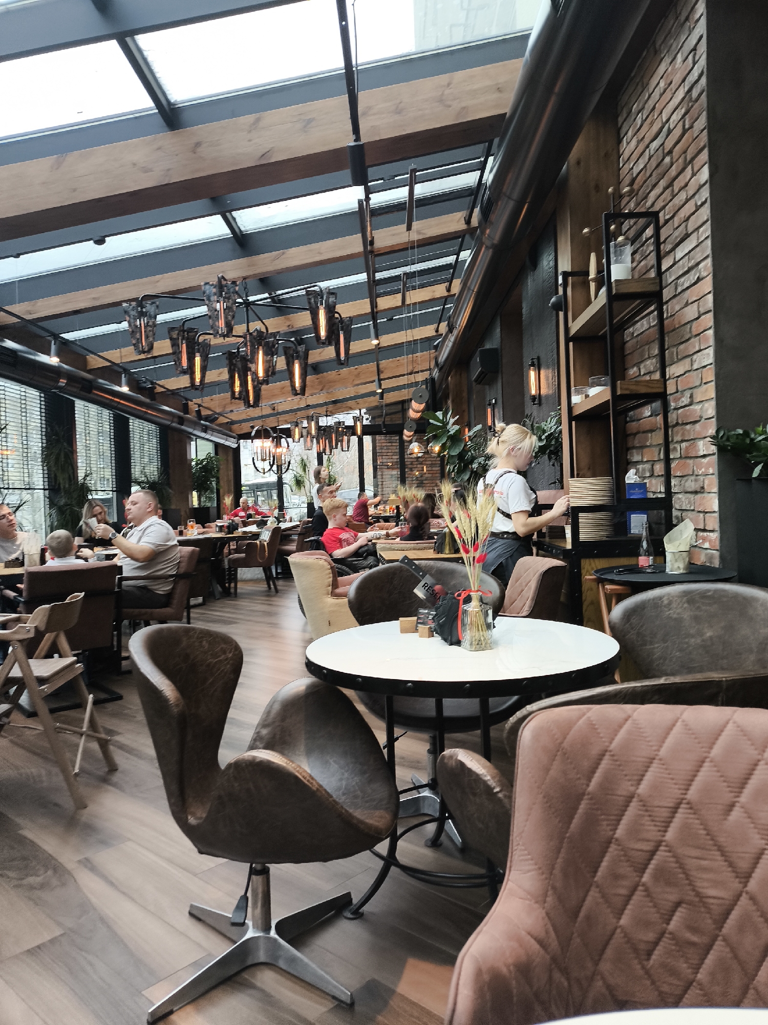 The image shows the interior of a restaurant. The restaurant features a glass roof and exposed wooden beams. Overhead are industrial-style light fixtures. A brick wall extends along one side of the restaurant. A woman with blonde hair is working near a shelf unit. There are several patrons at tables in the background. In the foreground, a round table with a floral arrangement stands surrounded by brown chairs. The location of the restaurant appears to be Izhevsk, Russia.