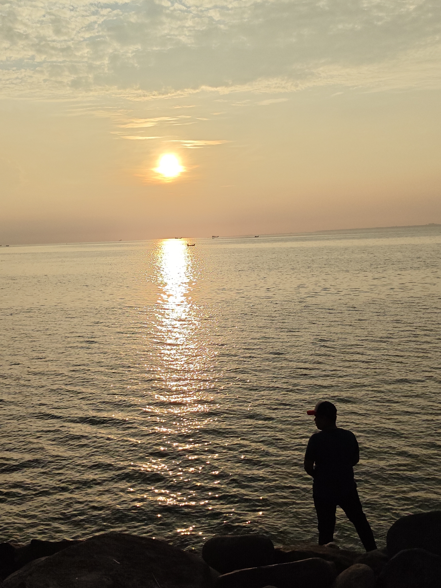 A lone figure wearing a dark t-shirt, dark pants, and a red baseball cap stands on a rocky shoreline, his gaze directed towards a sunset over the ocean. The location is likely in or near the city of Lhokseumawe, Indonesia, given the coastal environment and geographical details. The sun, partially obscured by clouds, casts a bright, shimmering path of light across the water's surface. Several small boats are visible on the horizon. The foreground is dominated by dark rocks, and the silhouette of the person is clearly defined against the bright water.