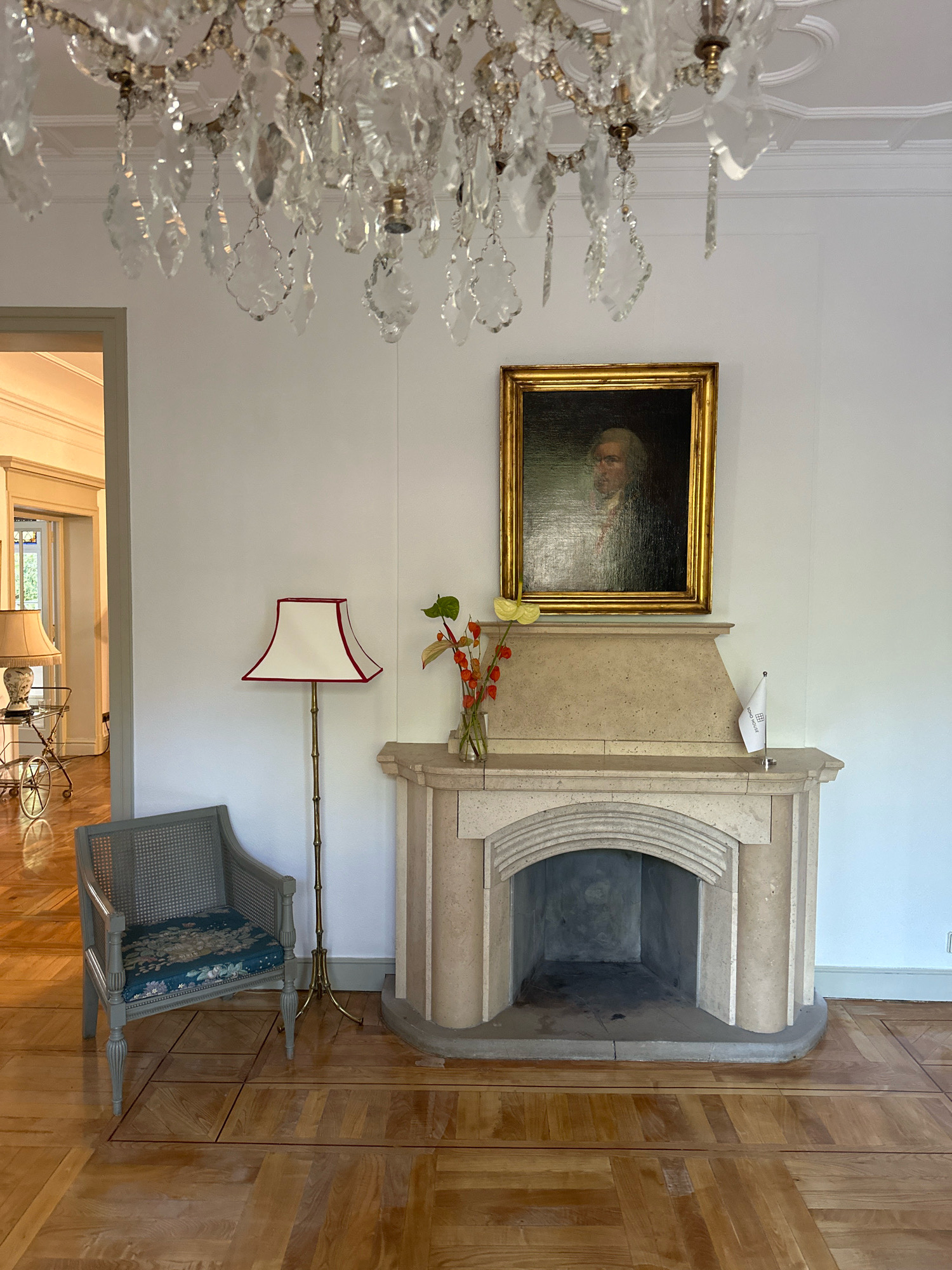 The image depicts a formal sitting room, likely within a historic building or upscale residence. Dominating the upper portion of the frame is an ornate crystal chandelier, its facets catching the light and casting a soft glow. Below, a classical-style fireplace with a stone surround is centrally located. Above the fireplace hangs a framed oil painting of a man in period attire. To the left of the fireplace, a light-colored armchair with a patterned cushion sits on a polished wooden floor laid in a parquet pattern. A tall floor lamp with a white shade and red trim stands beside the armchair. On the mantelpiece, a small flag on a stand is visible. The room appears to be illuminated by natural light entering from an adjacent area, visible through a doorway on the left, where another lamp can be seen on a trolley. The overall ambiance is one of refined elegance and historical character. There are no people present in the scene. The location is not specified as Madrid, Spain.
