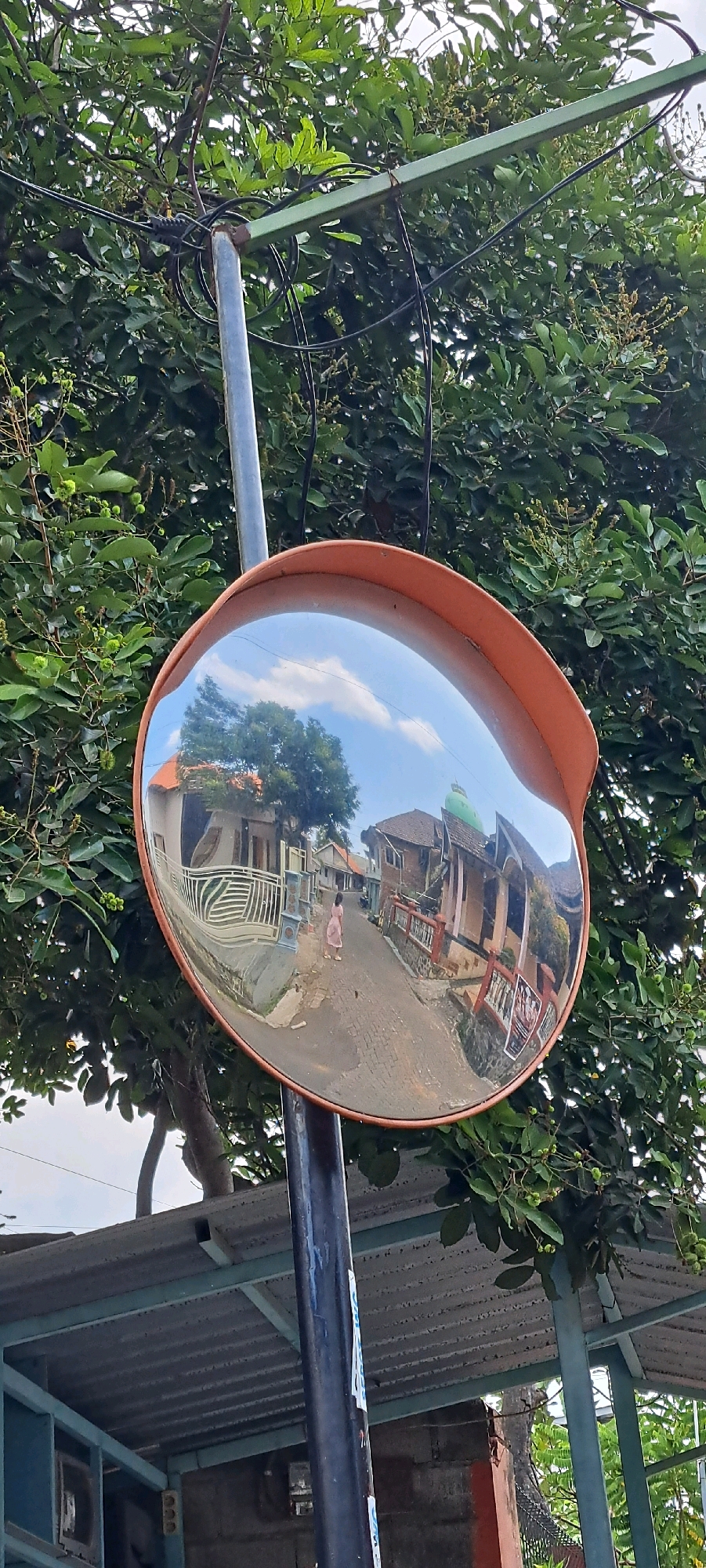 A convex traffic mirror is mounted on a metal pole, reflecting a street scene in Sumber Suko, Indonesia. The mirror shows a narrow, paved street lined with houses and buildings. A person, wearing a light-colored dress, is walking away from the viewer down the street. The sky with clouds is visible in the reflection. The mirror is surrounded by lush green foliage and branches of trees. The pole supporting the mirror is dark in color and appears to be metal. Below the mirror, a corrugated metal roof structure is visible, supported by metal beams. The overall environment suggests a residential area within a tropical or subtropical climate.