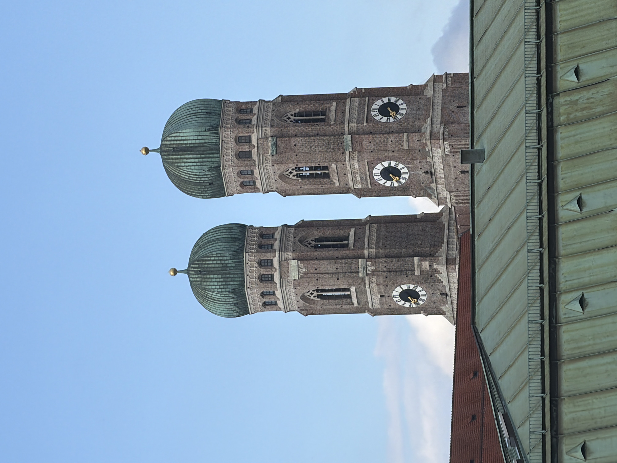 The image shows the twin towers of the Frauenkirche, a Roman Catholic cathedral in Munich, Germany. The towers are made of red brick and topped with green copper domes. Each tower has a clock face with Roman numerals. In the foreground, there are two overlapping copper roofs. The sky is a clear blue with a few wispy clouds.