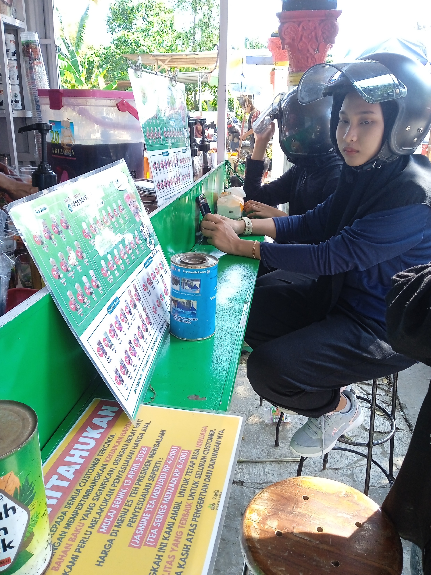 Two individuals wearing helmets sit at an outdoor stall in Kebondalem, Indonesia. The stall has a green countertop displaying menus and beverage options. One person is looking directly at the camera. The stall features an Arizona beverage dispenser and various canned goods. A sign details a price adjustment effective April 13, 2026, for "Es Teh Presiden" menu items, citing rising raw material costs. Both people appear to be sitting and waiting to order.
