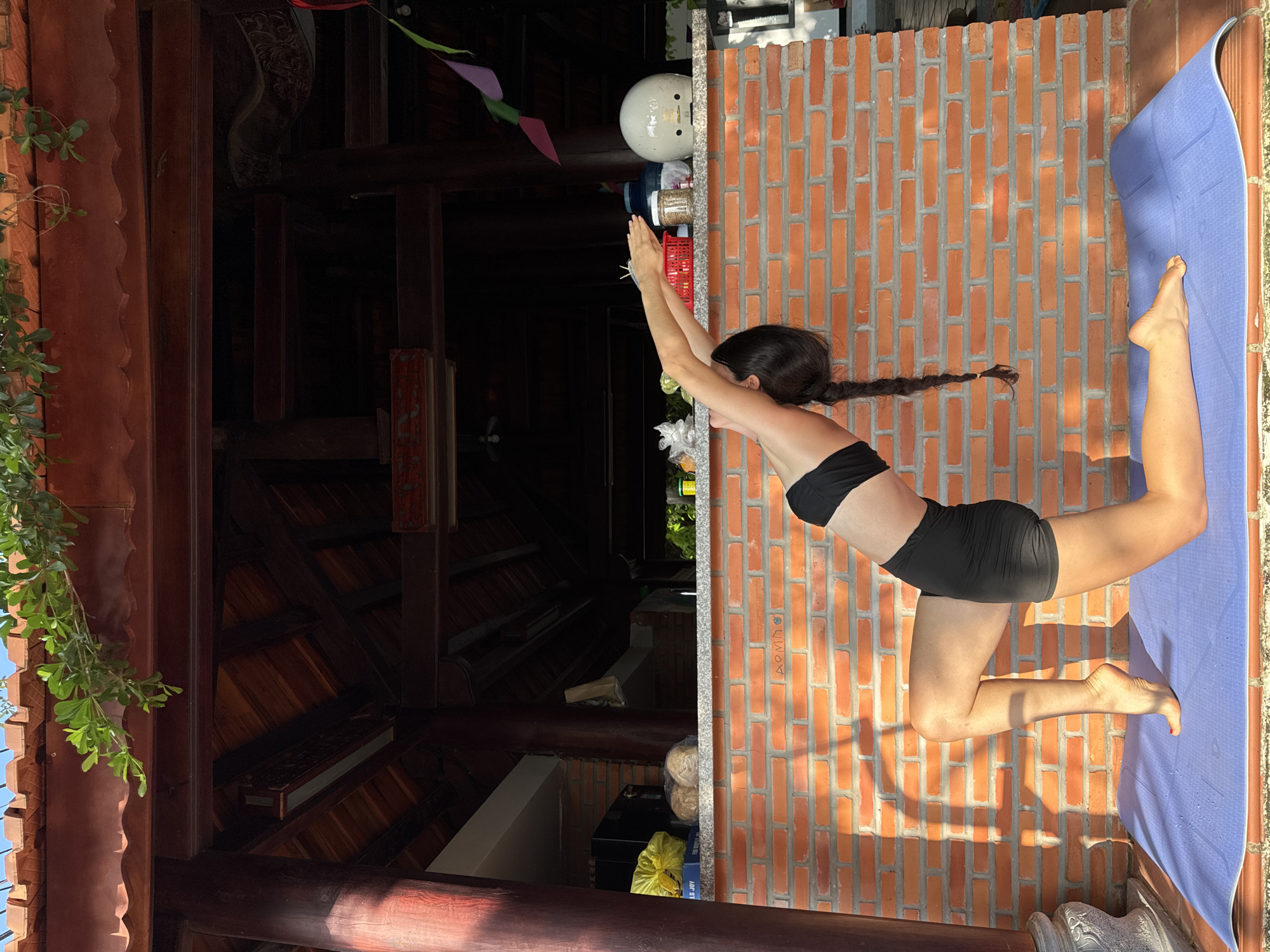 A long-haired individual, presumed female, in a black sports bra and black shorts, performs a kneeling backbend yoga pose on a purple mat. Her right knee is grounded, while her left foot is elevated on its toes. Her arms are extended overhead and backward, and her head is tilted back. The pose is executed on a reddish-brown tiled patio floor.
Behind the individual is a knee-to-hip-height wall constructed of orange-red bricks with white mortar, displaying angular sunlight shadows. A light grey speckled countertop rests on this brick wall. Visible items on the countertop, from left to right, include a yellow plastic bag, two packaged bread items, a red plastic basket, a clear jar containing light brown granular material, a large blue plastic jug, and a white motorcycle helmet placed upside down.
Behind the countertop, a dark wooden structure with pillars, beams, and a slatted ceiling indicates an open-air or semi-enclosed building. On the far left, a black appliance is partially visible. Multi-colored triangular flags (purple, pink, green) hang on the right side. Above the wooden structure, a traditional reddish-brown tiled roof is visible, with green foliage extending into the frame. A clear blue sky is visible above the roofline. This scene is consistent with architecture found in Mũi Né, Vietnam.
