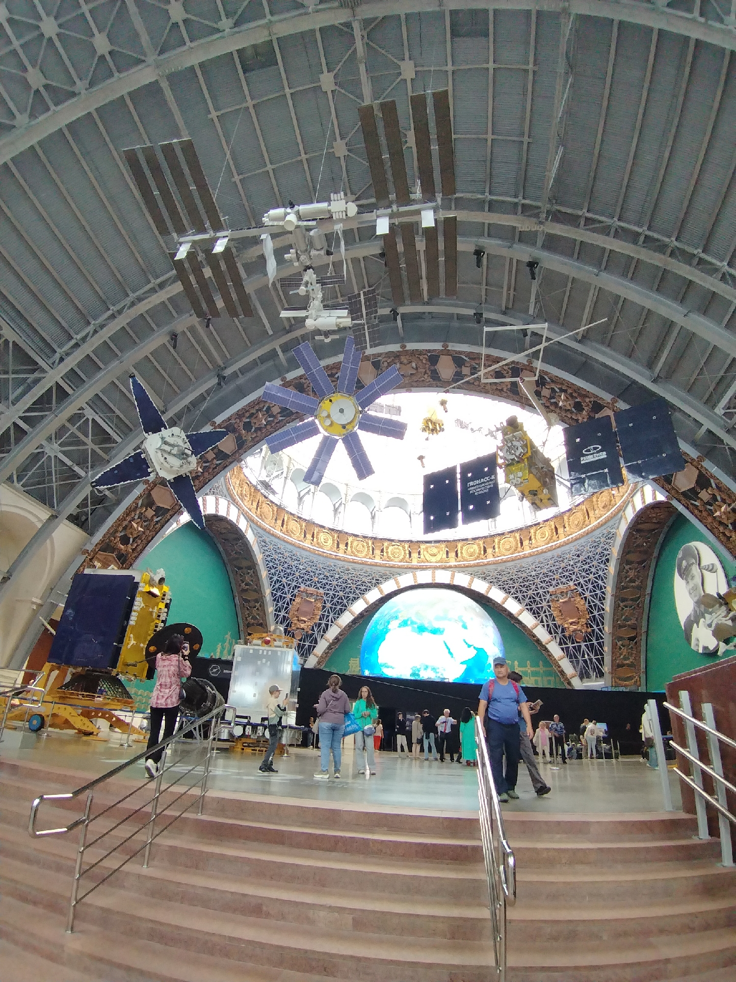 The image depicts the interior of the VDNKh exhibition center in Moscow, Russia, specifically the "Cosmos" pavilion (now known as the "Cosmonautics and Aviation" pavilion). The expansive, arched ceiling of the pavilion is visible, supported by a complex metal framework. Hanging from the ceiling are several large-scale models of spacecraft and satellites, including what appears to be a model of the International Space Station in the upper left. A prominent spherical display showing the Earth is visible in the background.
In the foreground, a wide staircase with metal handrails leads down into the main exhibition area. Several individuals are present on the stairs and on the floor below. On the stairs, a person in a pink and white patterned shirt is walking up, holding a phone and appearing to take a photo. Further down the stairs and on the floor, various visitors are seen walking around and observing the exhibits. A man in a blue t-shirt and dark pants is descending the stairs, and another person in a grey shirt and jeans is also on the stairs.
The exhibition space features teal-colored walls with decorative golden accents and what appear to be murals or displays depicting space-related themes. Various exhibits, including models of rockets and satellites, are positioned throughout the space. The overall impression is that of a public exhibition hall dedicated to space exploration.