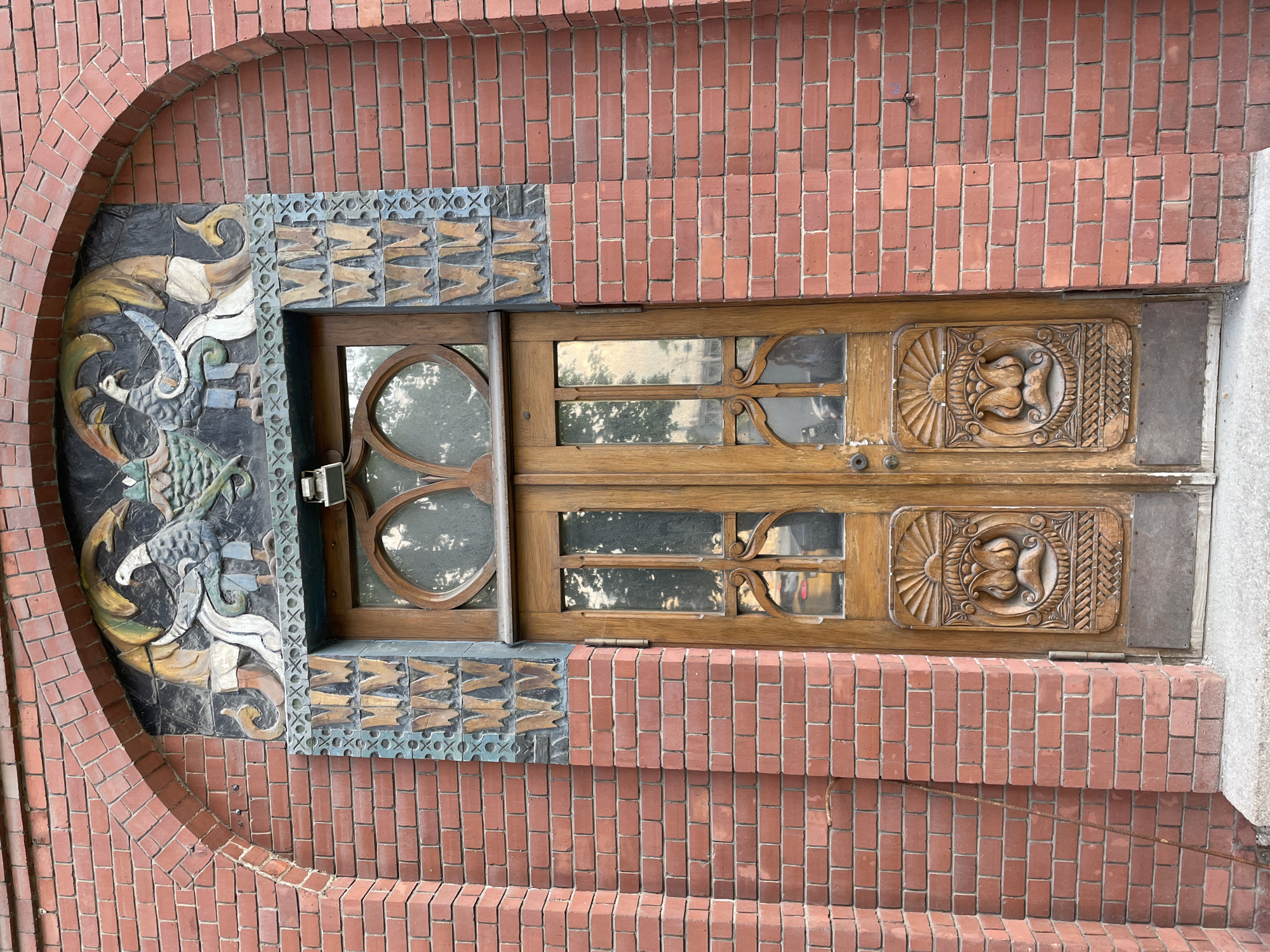 The image shows the facade of a building in Moscow, Russia, featuring an ornate wooden door set into a brick wall. The door is flanked by decorative tiles with a dragon motif on one side. The brickwork forms an arch around the left side of the door frame. The door is made of wood with carved details, and contains windows. The background suggests an urban environment with trees reflected in the windows.