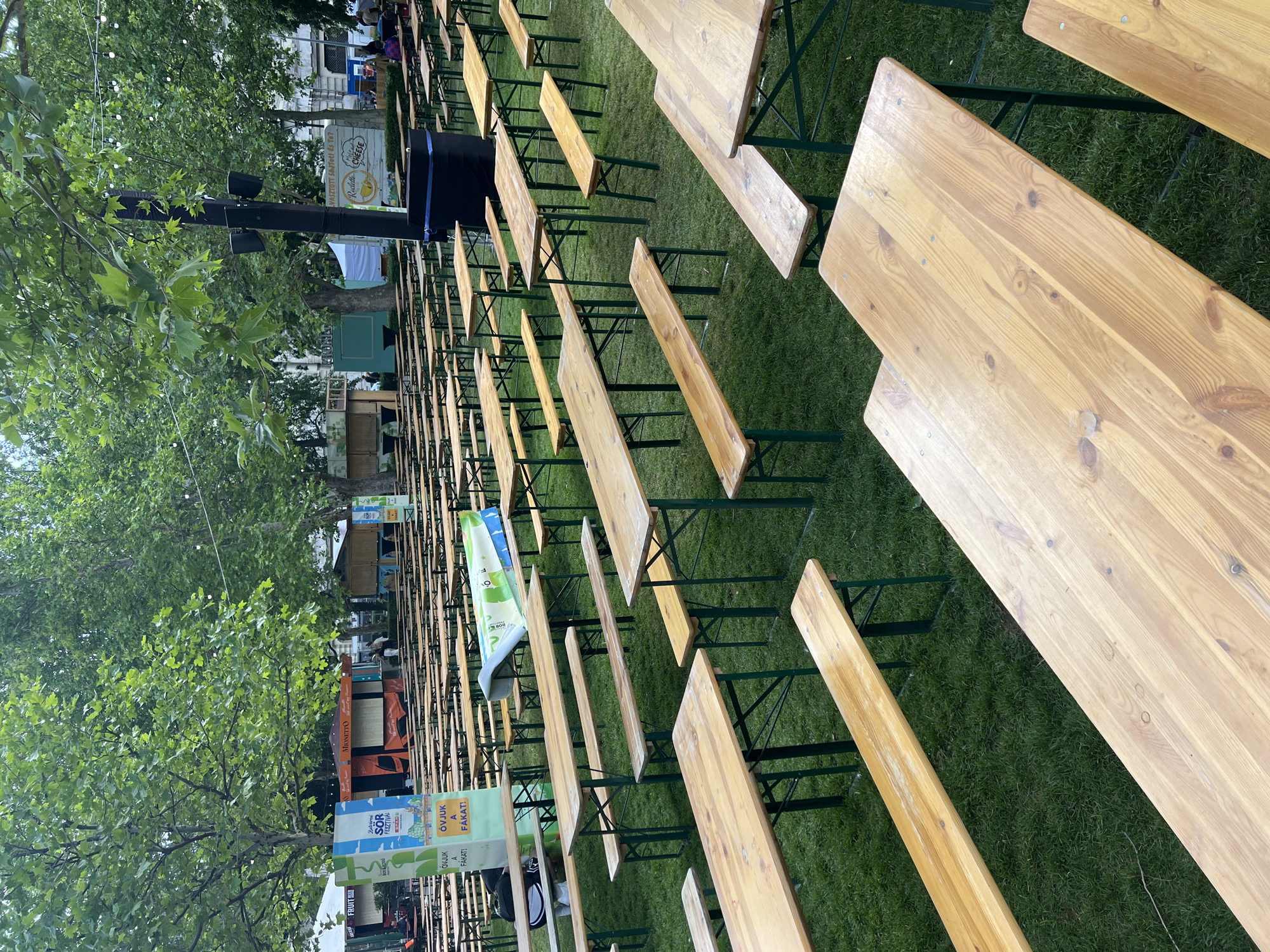 The image shows rows of wooden tables and benches set up on a grassy area. The rows extend into the distance, suggesting a large event or gathering space. In the background, there are trees, booths, and signs, some of which have partially visible text in Hungarian, including "SÖR FESZTIVÁL" (Beer Festival) and "ÓVJUK A FÁKAT!" (Let's protect the trees!). Another sign mentions "Redette CHEESE". The scene suggests an outdoor event or festival in a park-like setting in Budapest, Hungary.