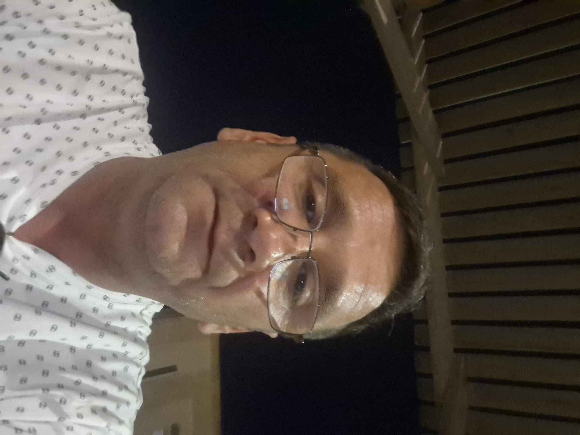 A man wearing glasses and a white shirt with a dark pattern is pictured from the chest up. He appears to be outdoors at night, with a dark background that suggests it is nighttime. Above him, a structure made of wooden slats is visible, possibly a pergola or a roof. The lighting is dim, and the man's face is partially illuminated, casting shadows. The image appears to be a selfie, as the man is looking directly at the camera. The overall scene suggests a casual, relaxed setting, possibly a backyard or patio area. There is no information to indicate the location in Yerocham, Israel.