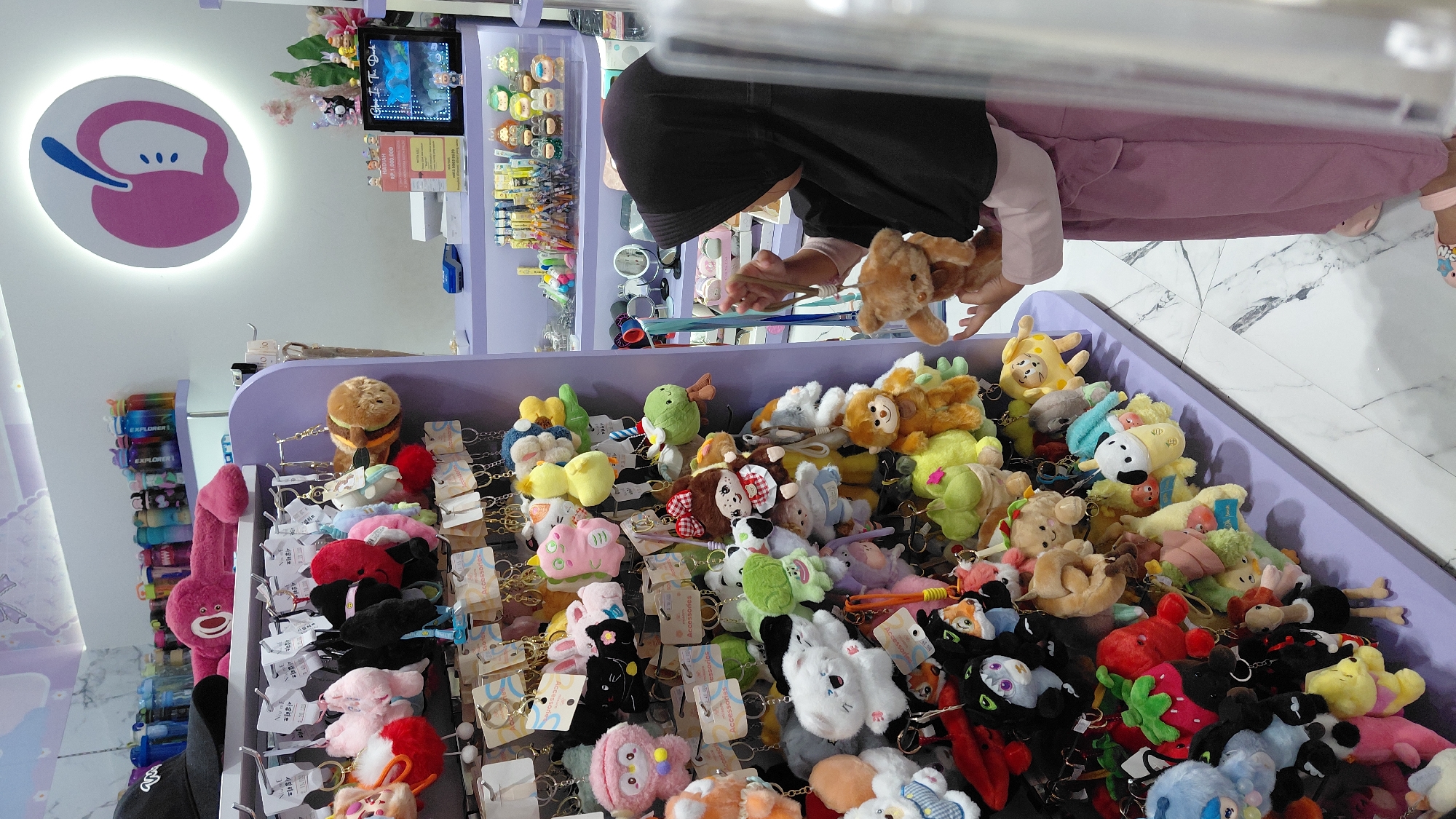 In a retail store, possibly in Purwokerto, Indonesia, a young girl wearing a black hijab and a pink outfit stands on the right side of the frame. She is holding a small, light brown teddy bear keychain in her right hand, examining it. In front of her is a vertical display rack filled with numerous colorful plush keychains, including cartoon characters, animals, and food items. The keychains are predominantly in pastel colors like pink, yellow, green, and blue. Behind the girl, shelves are stocked with more merchandise, including bottles and other small items. The store has a bright, cheerful ambiance with purple display fixtures and a marble-patterned floor. A glowing pink apple logo is visible on the wall in the upper right corner of the image.