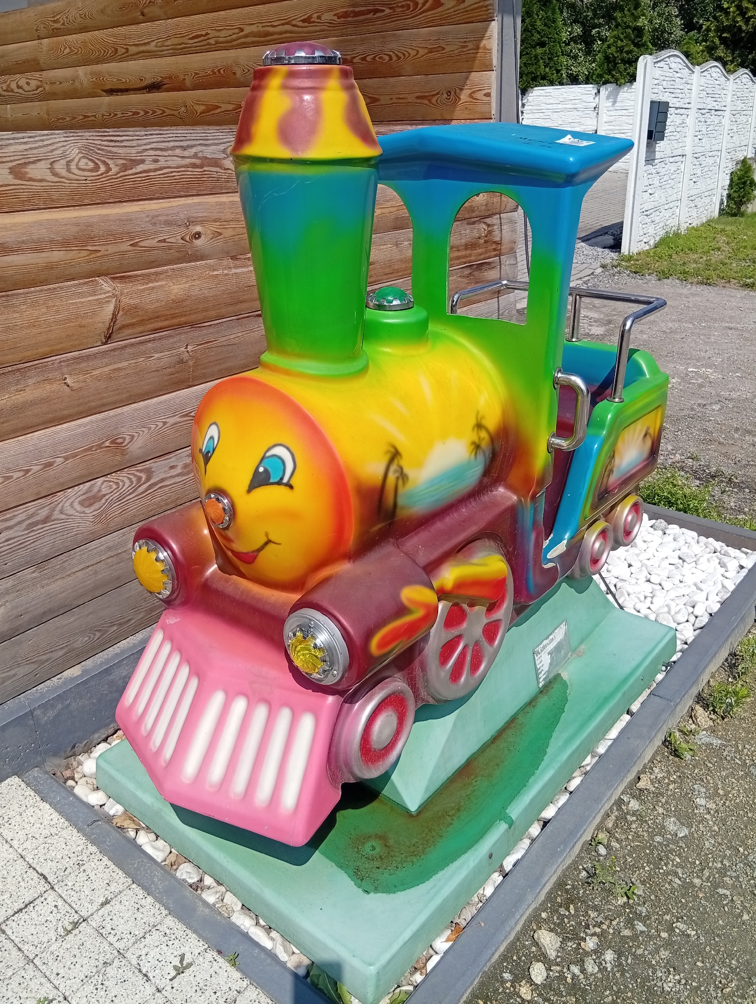 A brightly colored, toy train ride is situated outdoors in Brzezia Łąka, Poland. The train is primarily yellow and green with accents of blue, pink, and red, and features a cartoonish face with blue eyes and a smiling mouth. The train is positioned on a light green base, surrounded by white decorative stones. In the background, a wooden wall is visible to the left, and a white, decorative fence with greenery behind it is to the right. The ground is a mix of gravel and paving stones. The scene appears to be a sunny day.
