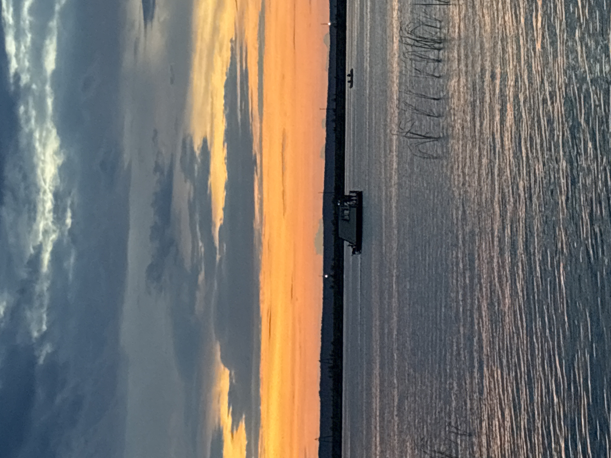 The image captures a tranquil sunset scene over a body of water, likely the Kama River, in Chaikovsky, Russia. A large, flat-bottomed boat, possibly a barge or ferry, is positioned in the middle ground, with several figures visible on its deck. To the right, a smaller boat with two occupants glides across the water. The foreground is dominated by rippling water with reeds or tall grass emerging from the surface. The horizon is marked by a dark, silhouetted treeline, punctuated by industrial structures and lights. The sky is a dramatic display of color, with vibrant orange and yellow hues from the setting sun blending into darker, stormy blue and grey clouds. The overall impression is one of peacefulness and the beauty of nature.