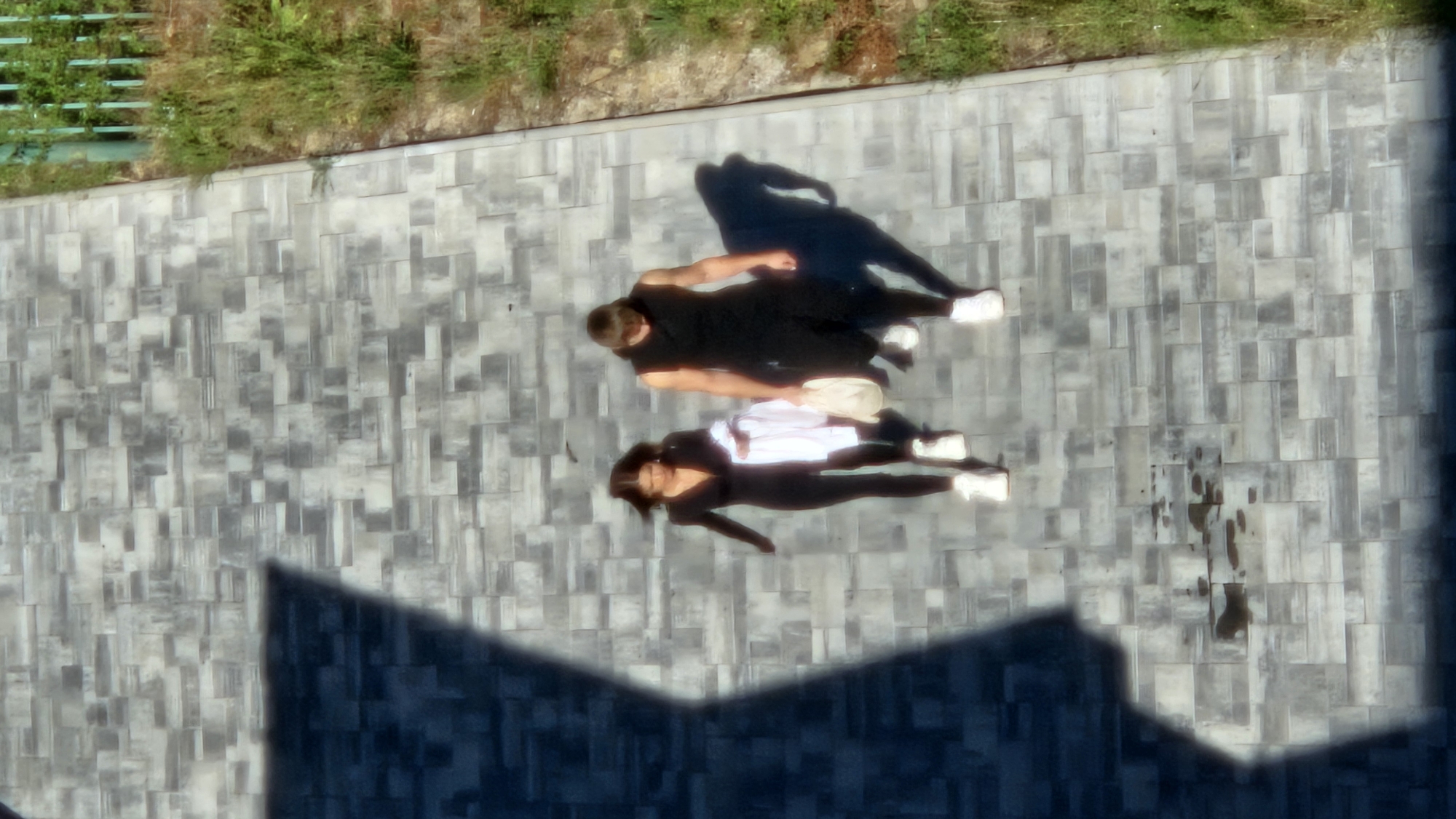 An overhead, slightly blurry shot captures two individuals walking side-by-side on a grey tiled pathway. The person on the left is a woman wearing a black catsuit and white sneakers, carrying a folded white garment. The person on the right is a man wearing a black sleeveless top, dark pants, and white sneakers, carrying a light-colored bag. They are walking away from the viewer, casting shadows on the textured tiles. The pathway is bordered on the right by overgrown green grass and a partial view of a green metal fence. A large, dark shadow, likely from a building, occupies the left side of the frame, partially obscuring the pathway. The scene appears to be an outdoor public space, possibly a park or walkway, in the city of Prešov, Slovakia.