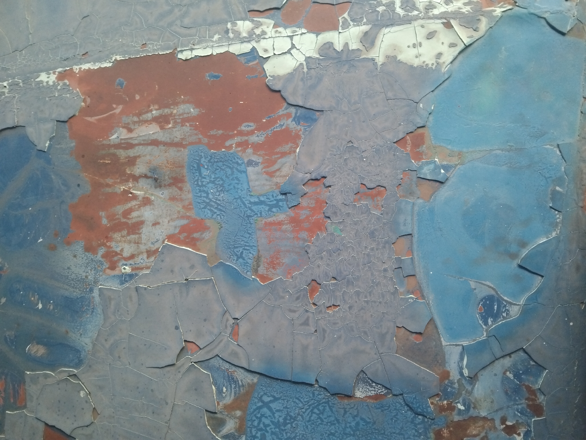This is a close-up, abstract image of peeling paint on a surface. The paint is layered, with visible cracks and chips revealing underlying colors. The dominant colors are shades of blue, reddish-brown, and grey, with some white peeking through. The texture is rough and uneven due to the distressed paint. There are no discernible objects, people, or actions, and the setting is abstract and lacks specific environmental cues. Therefore, it is not possible to provide a description of this image that includes the location in the city of Tempeh, Indonesia, as no such information can be inferred.