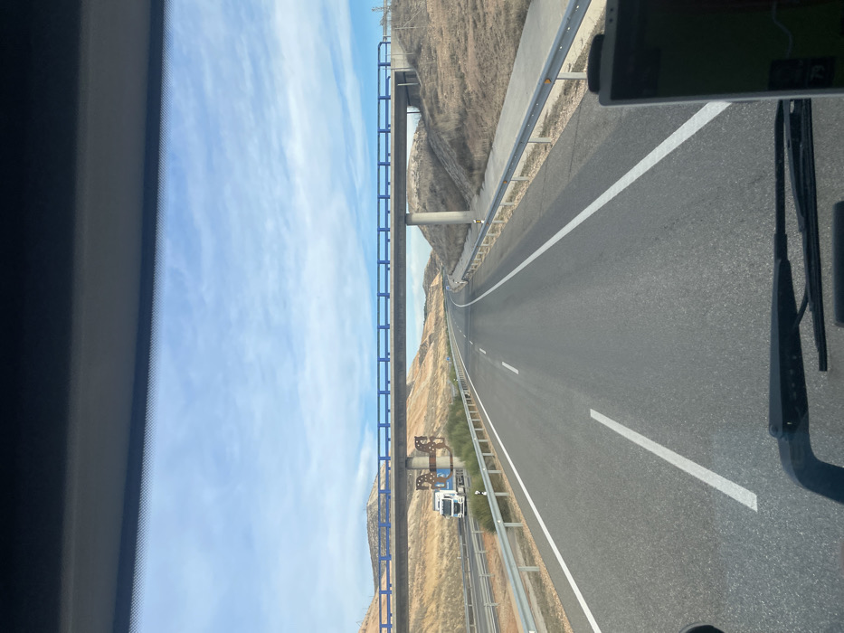 A highway stretches forward, flanked by a rugged, arid landscape. An overpass bridge with blue railings is on the left. A white truck appears under the bridge. A cloudy blue sky is above the landscape. The perspective is from inside a vehicle with windshield wipers visible. The image is rotated counterclockwise.