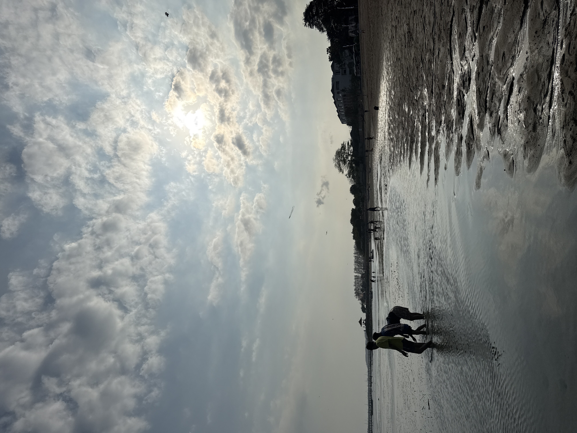 The image depicts a seaside location, likely a beach, in Denpasar, Indonesia, under a cloudy sky with the sun partially obscured. The foreground features shallow water with ripples reflecting the sky and the sandy shore. Several figures, silhouetted against the light, are visible. In the immediate foreground, three individuals are wading through the water, two of them appearing to bend down. Further in the distance, along the shoreline and in the water, other people are scattered, some walking and others potentially engaging in activities. The background reveals a line of trees and buildings along the coast, with a structure that appears to be a gazebo or pavilion near the water's edge. The overall atmosphere is serene and suggests a late afternoon or early evening setting.