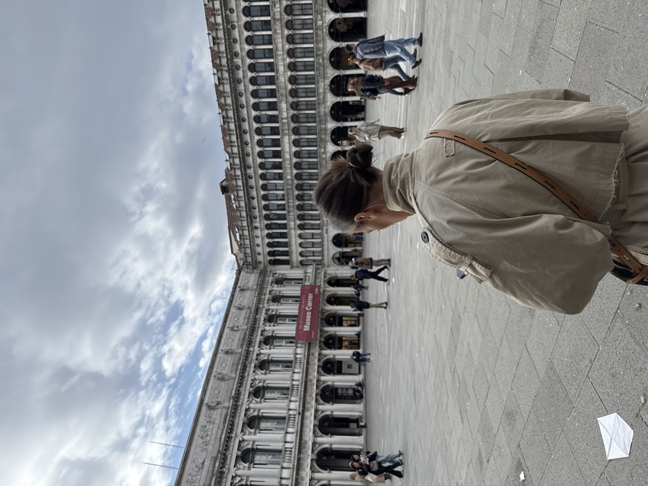 The image depicts a paved urban square in Venice, Italy, identified as Piazza San Marco, with the Museo Correr visible on a building in the background. The sky is largely overcast with patches of blue.
In the immediate foreground, facing away from the camera, is a person with dark hair styled in a messy bun, wearing a light beige or khaki-colored jacket and a brown strap (likely from a bag) across their back. The individual appears to be holding a smartphone in their left hand, which is partially visible.
The square itself is paved with large, light-colored flagstones. A crumpled piece of white paper or litter lies on the ground in the lower-left quadrant.
In the mid-ground and background, multiple individuals are seen walking across the square and near the surrounding buildings. The prominent building on the left features neoclassical architecture with multiple arched windows, pilasters, and a decorative cornice. A red banner displaying "Museo Correr" in white text is affixed to its facade. To the right, another large building with a long series of arched windows and colonnades extends into the distance.