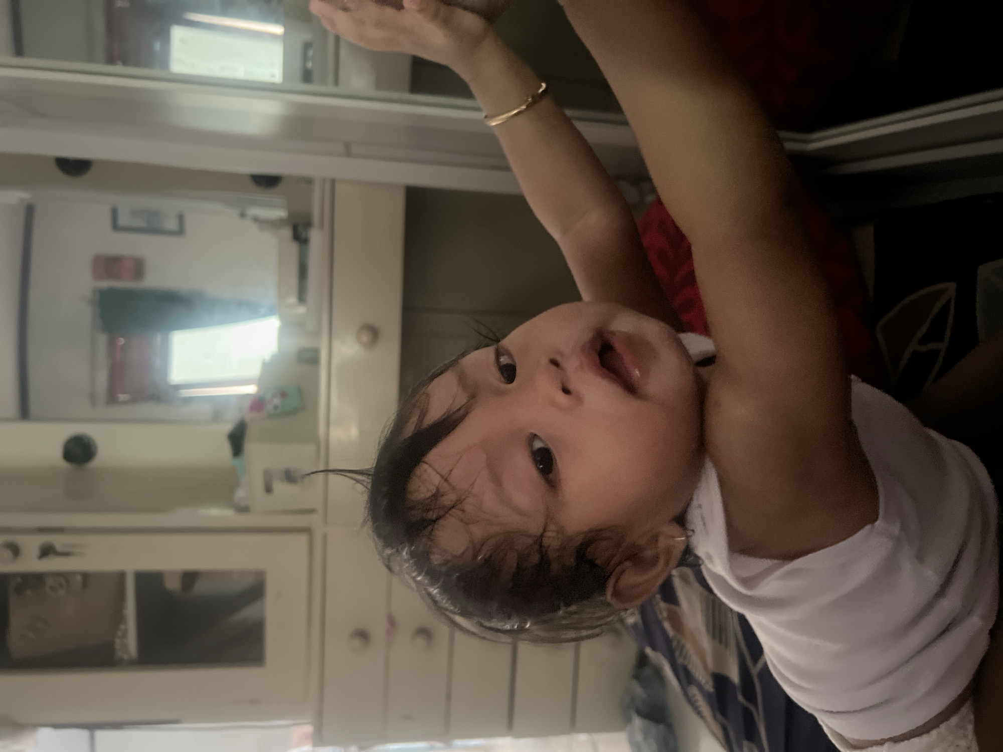 A child, likely an infant, is shown close-up in an indoor setting. The child is wearing a white shirt. The location appears to be a home in Kecamatan Pati, Indonesia, based on contextual information. In the background is what appears to be cabinetry, potentially in a kitchen or bathroom. One arm is raised. A bracelet is visible on the wrist of the arm.