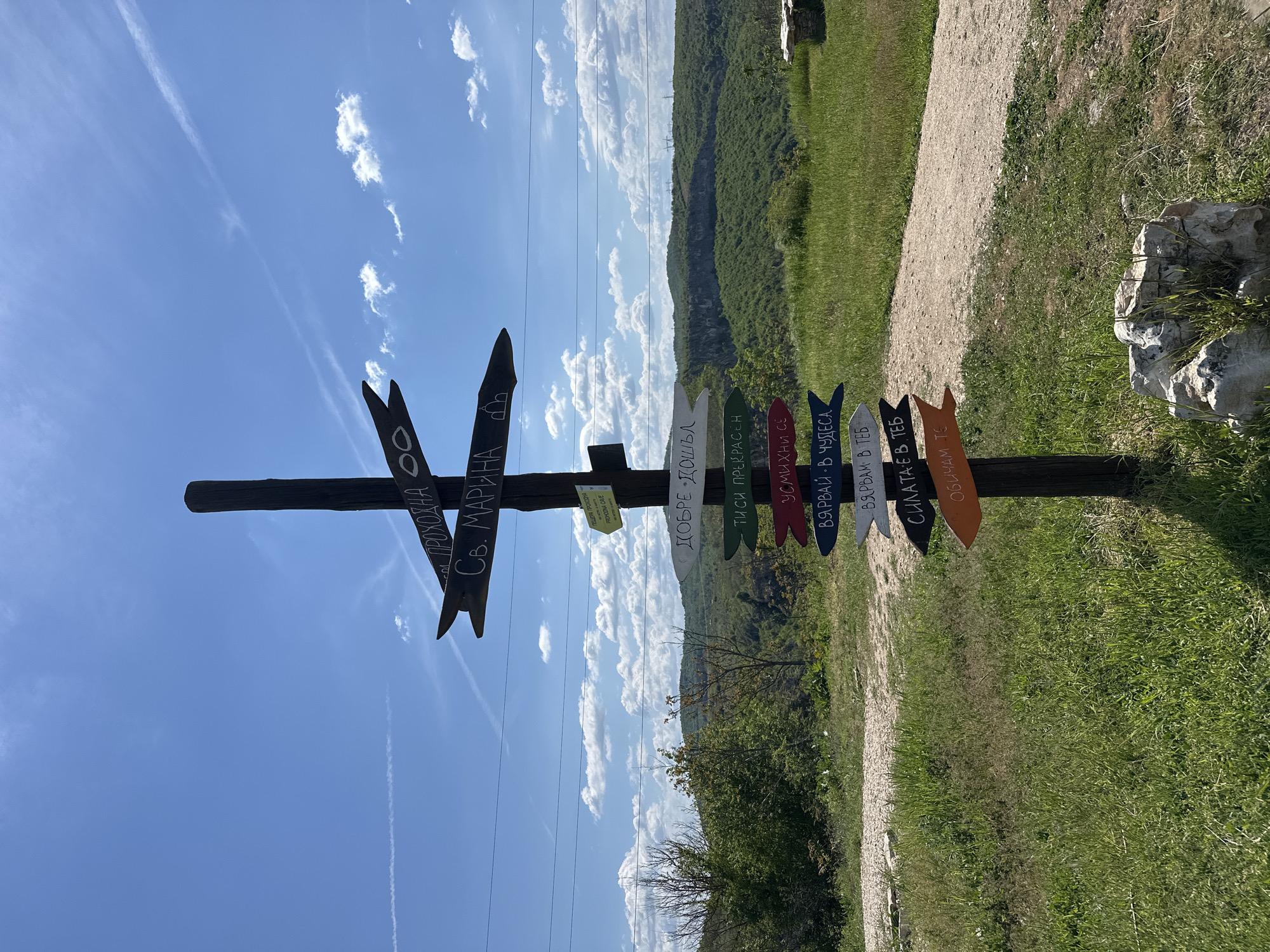 The image shows a direction sign post in a rural location. The post is brown and weathered, with several smaller, arrow-shaped signs attached, each with a different inscription written in Cyrillic. The post has two black arrow-shaped signs at the top. Behind the post is a dirt path bordered by green grass, leading towards a lush, green hillside. The sky is blue with scattered clouds, and some power lines are visible. The Cyrillic text on the sign includes phrases like "СВ. МАРИНА" "ПРОХОДНА", "ПЕЩЕРА ПРОХОДНА PROHODNA CAVE" "ДОБРЕ ДОШЪЛ", "ТИСИ ПРЕКРАСЕН", "УСМИХНИ СЕ", "ВЯРВАЙ - В ЧУДЕСА", "ВЯРВАМ В ТЕБ", "СИЛАТА Е В ТЕБ", "ОБИЧАМ ТЕ". The overall scene suggests an outdoor area near a tourist attraction, likely in or near Karlukovo, Bulgaria, as the sign references Prohodna Cave, a known landmark there.