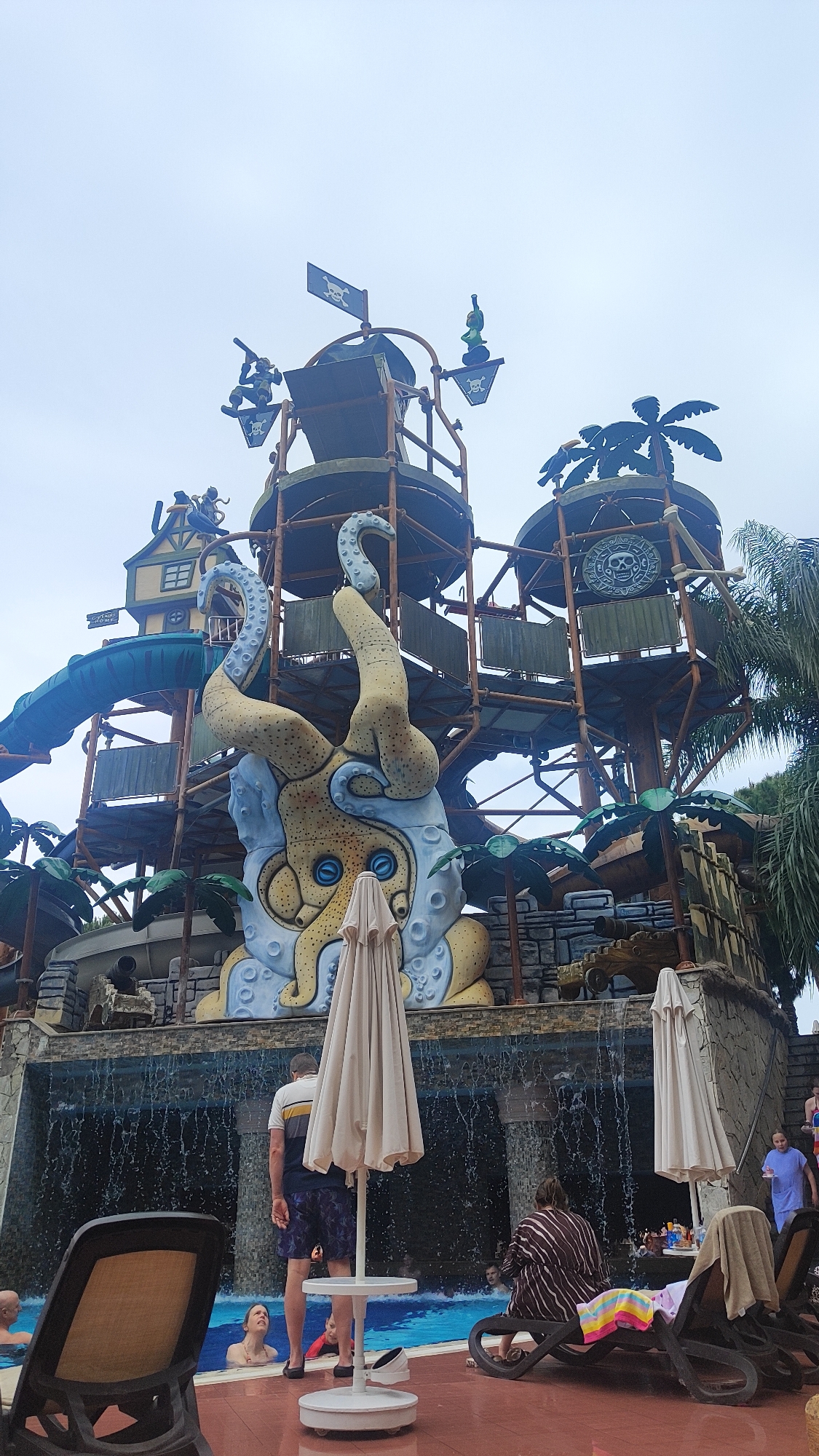 The image depicts a vibrant waterpark scene, specifically at what appears to be Water Planet Aqua Park in Alanya, Turkey. The central focus is a massive, multi-tiered water play structure dominated by a large, light yellow and light blue octopus (Kraken) sculpture with prominent tentacles and eyes. This sculpture forms part of the structure's base, which is further adorned with faux rockwork and tropical palm trees.
Above the octopus, the structure ascends with multiple levels of platforms, featuring a pirate theme. Notable elements include black flags with white skull and crossbones (Jolly Roger), figures resembling pirates (one holding a musket), a small building resembling a pirate's shack, and large circular emblems with skull motifs. Green and blue water slides snake down from various levels. At the very top, another pirate flag flies, and what appears to be a large water-dumping bucket is visible.
At the base of the structure, a man-made waterfall cascades into a swimming pool. In the foreground, surrounding the pool, is a tiled area with several brown lounge chairs, some occupied, and multiple beige closed parasols. A white table stands next to one parasol.
People are visible in and around the pool. Inside the pool, several individuals are partially submerged, including a child in red swimwear. On the poolside, a male subject, viewed from the back, wears a white, blue, and black striped shirt with patterned shorts, standing near the pool's edge. A female subject, also viewed from the back, is seated on a lounge chair, wearing a striped brown and white garment. Other individuals are partially visible in the background near additional lounge chairs. The sky above is overcast and light grey.