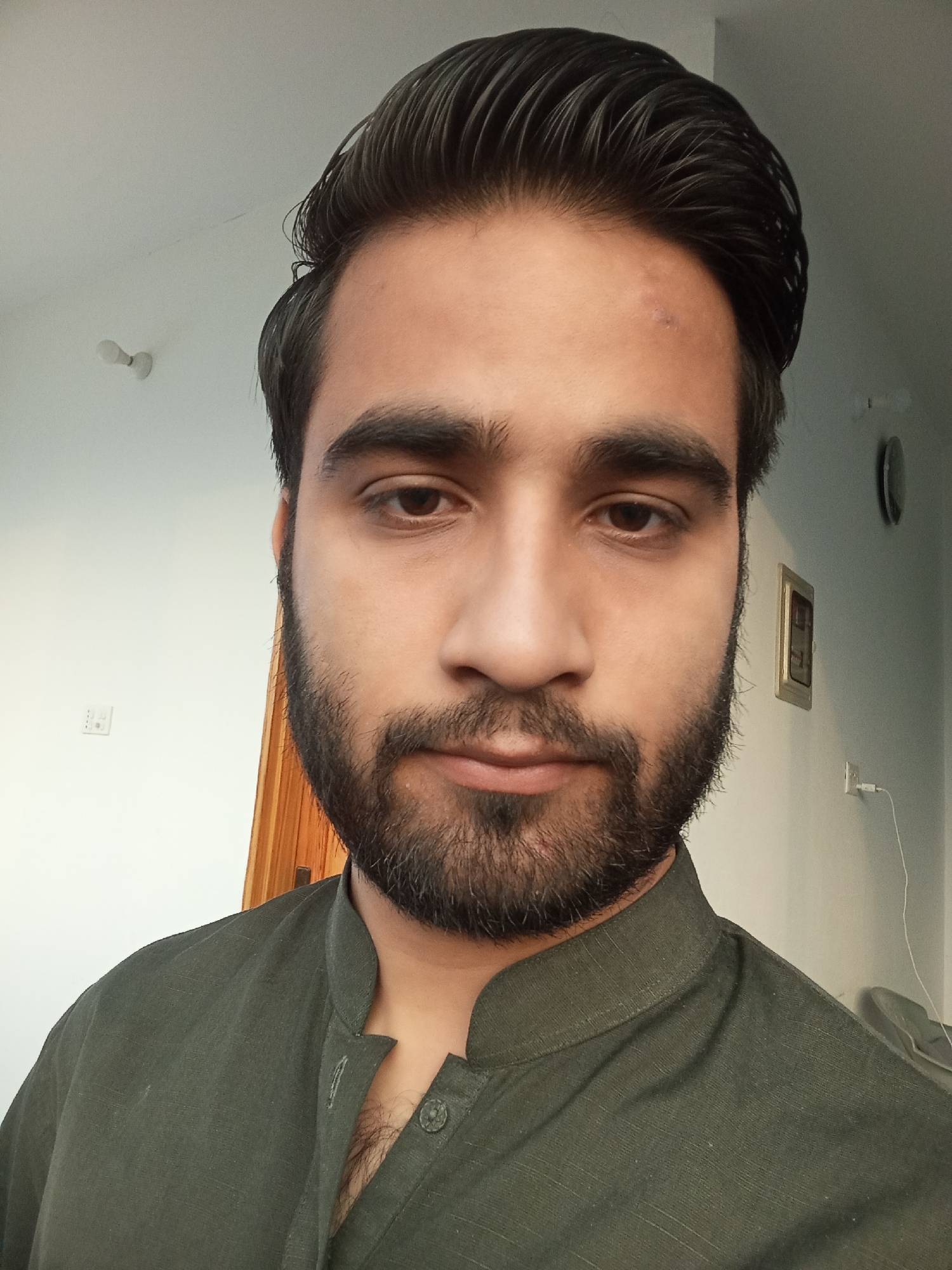 The media file is a close-up, front-facing selfie of a young man with dark hair and a beard. He is wearing a dark green, collared shirt. The background is out of focus and appears to be an indoor setting, with a white wall and a wooden door visible. There is a light fixture on the ceiling and what looks like an electrical outlet on the wall. The lighting suggests it is daytime. The man's expression is neutral. There is no visible text in the image.