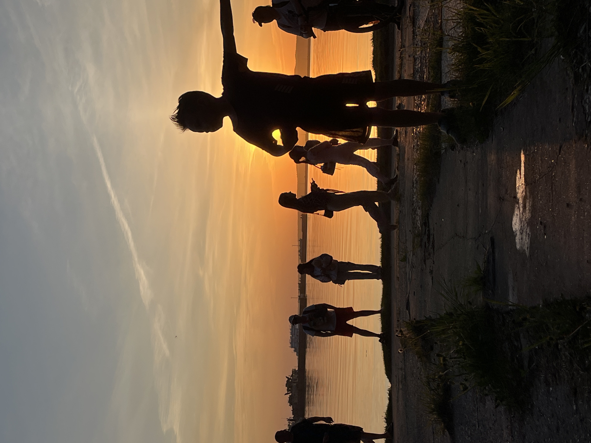 Several silhouetted people are standing on a concrete platform next to a body of water at sunset. The sky is a gradient of yellow and orange near the horizon. A vapor trail is visible in the sky. Tall grass and small puddles are at the edge of the platform.