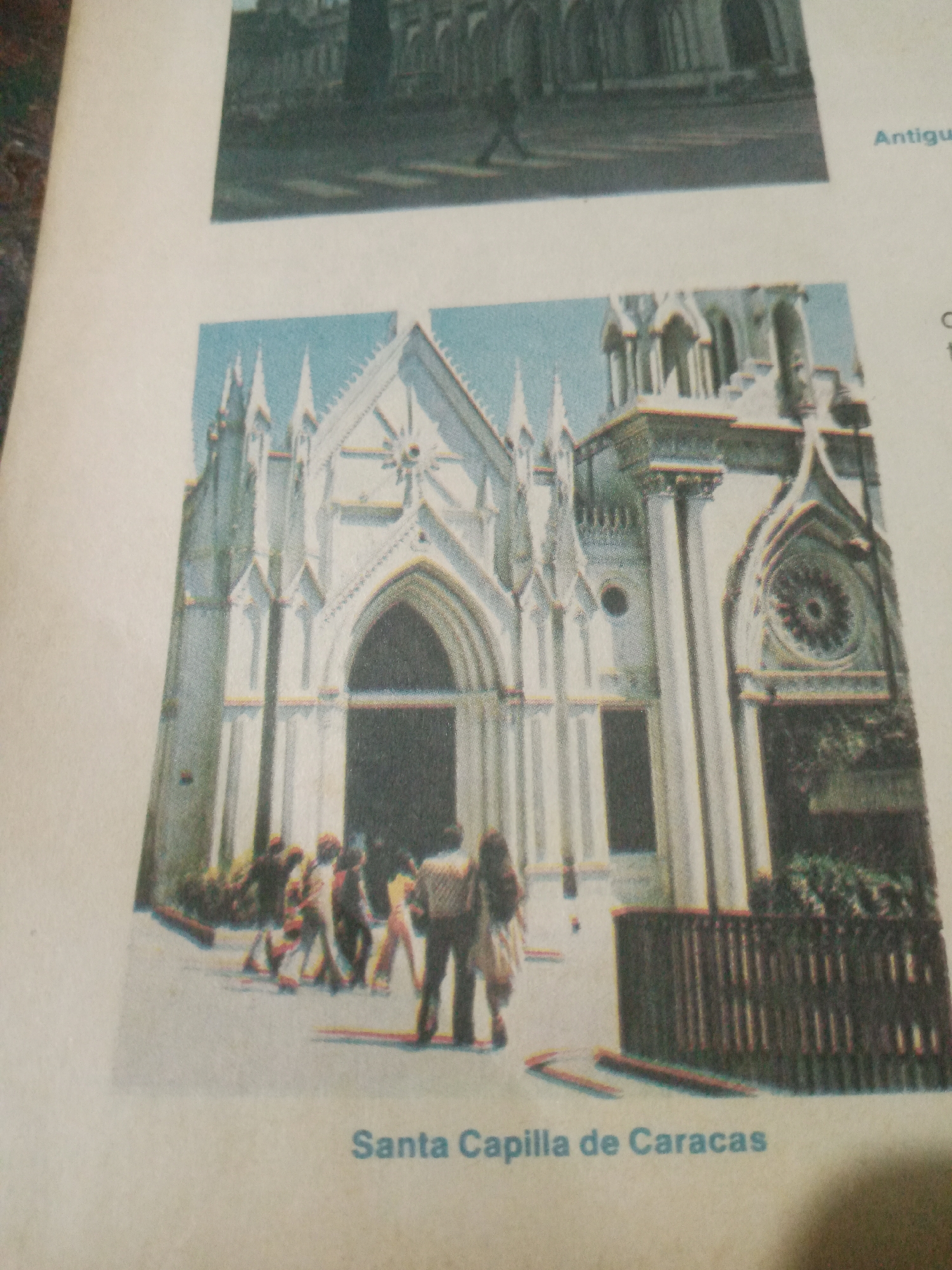 The image depicts the Santa Capilla in Caracas, Venezuela. The facade of the building is primarily white with Gothic architectural elements, including pointed arches and spires. A large, arched entrance is visible in the center, flanked by columns and ornate detailing. A rose window is present on the right side of the building. In front of the chapel, a group of people, some walking and some standing, are gathered. The ground appears to be paved. A dark wooden fence or railing is located to the right of the group and in front of some greenery. The text "Santa Capilla de Caracas" is printed below the image, identifying the subject.