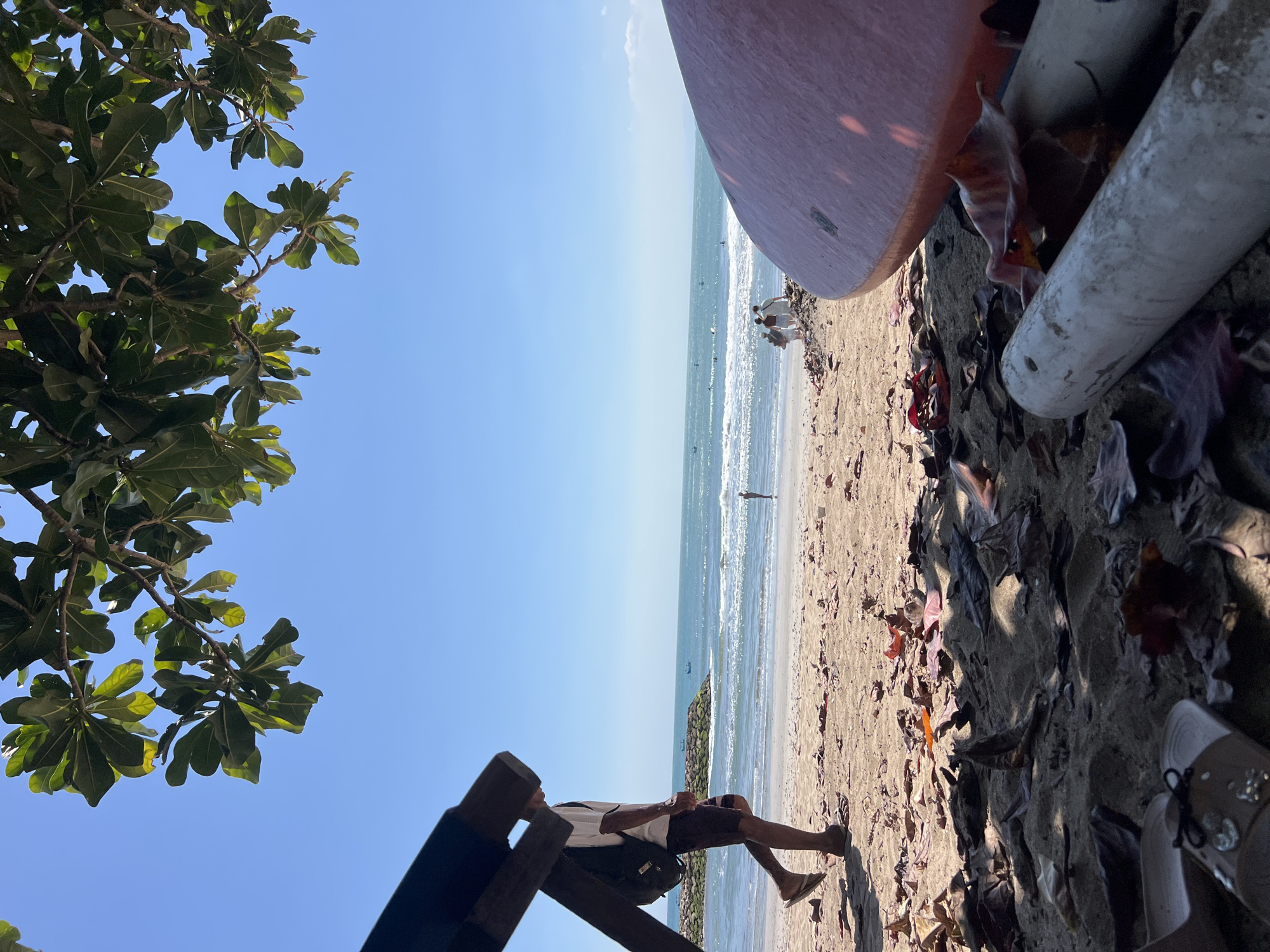 A man wearing a white t-shirt, dark patterned shorts, and flip-flops walks on a sandy beach in Kuta, Indonesia, carrying a backpack. He is partially obscured by a dark wooden structure. In the background, the turquoise ocean meets a clear blue sky. Several boats are visible on the water, and a person stands near the shore. Further in the distance, a group of people walks along the beach. In the foreground, a large, rounded orange object, likely a surfboard, lies on the sand. Scattered dry leaves and two cylindrical white objects are also present in the foreground. The scene appears to be a bright, sunny day.